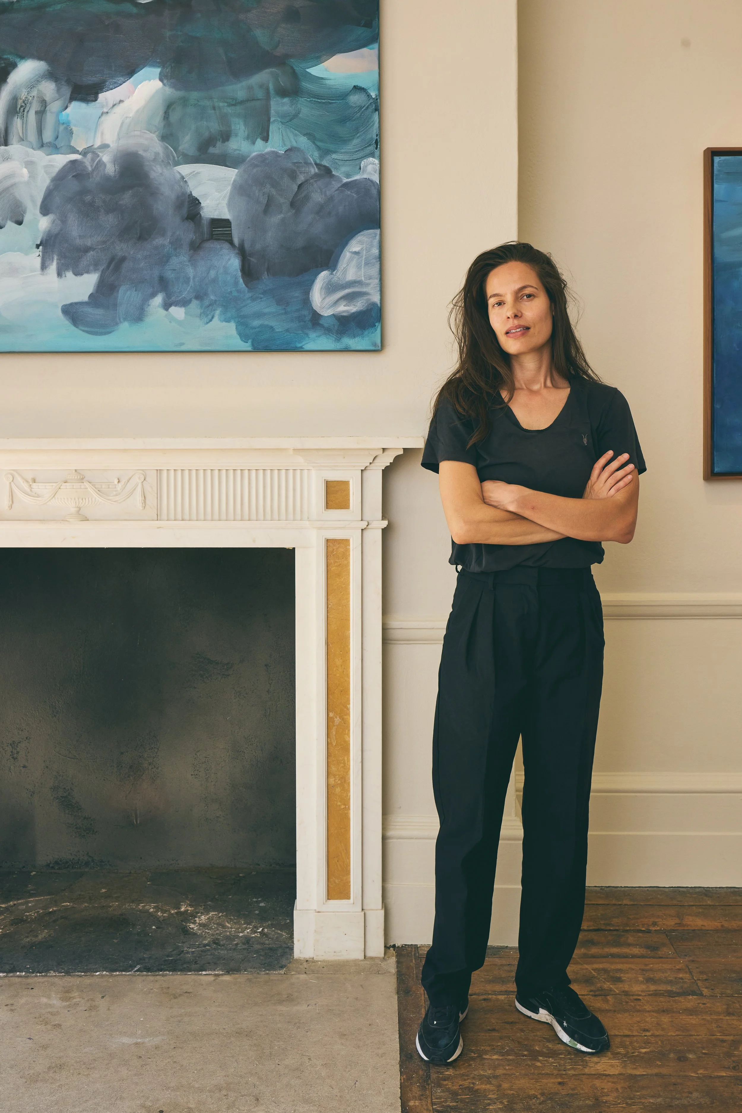 A woman with long dark hair, wearing black clothing and black sneakers, standing with arms crossed in an art gallery near a fireplace, with abstract blue and gray paintings on the wall behind her.