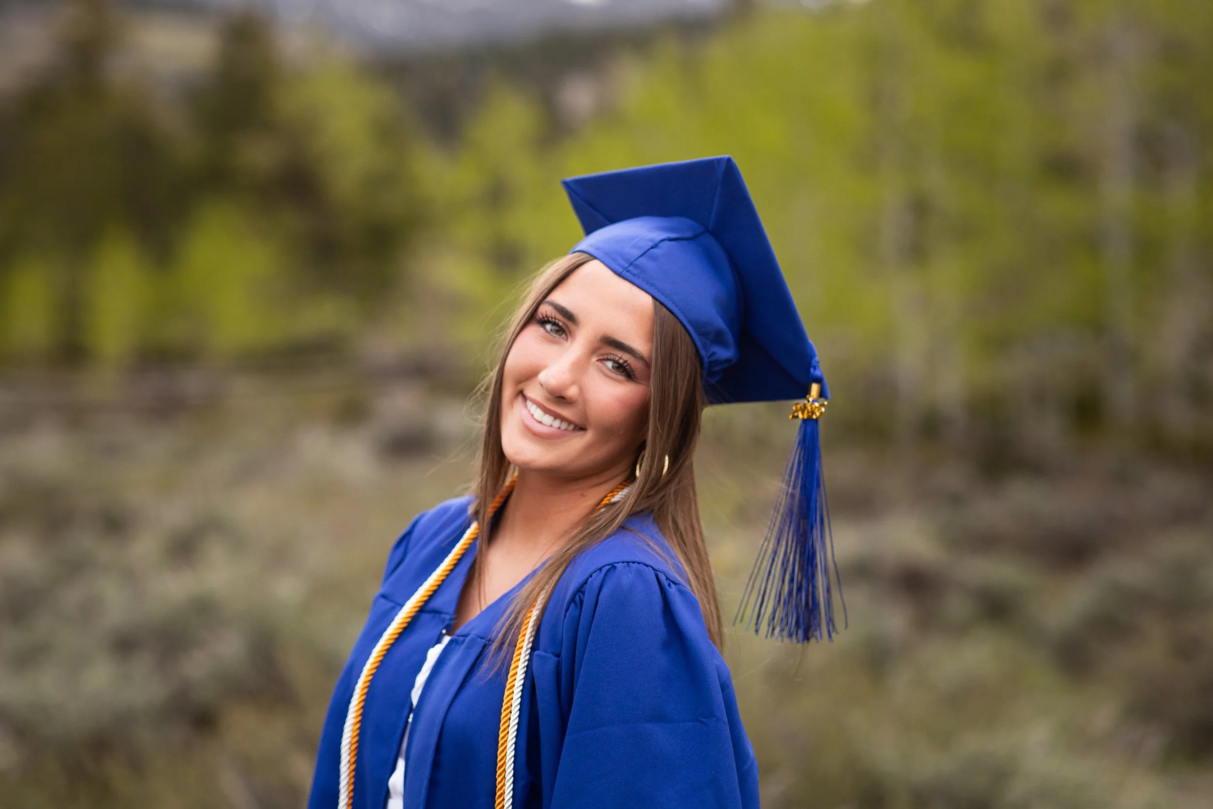 A young woman in a sleeveless yellow dress with black floral patterns standing outdoors by a wooden fence, with autumn foliage and mountains in the background.
