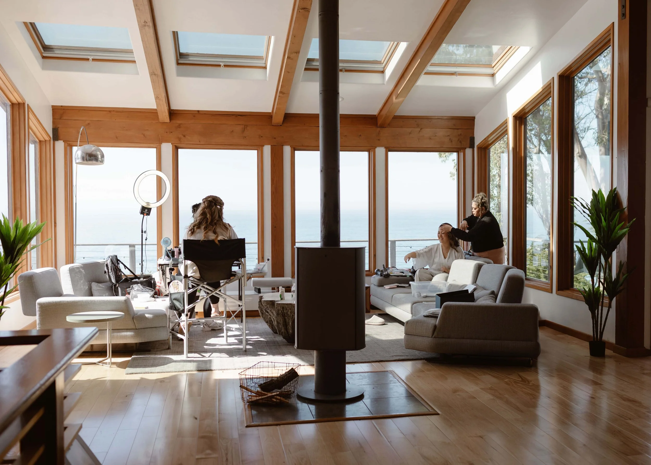 Bride getting hair and makeup done inside a bright coastal Airbnb with large ocean-facing windows.