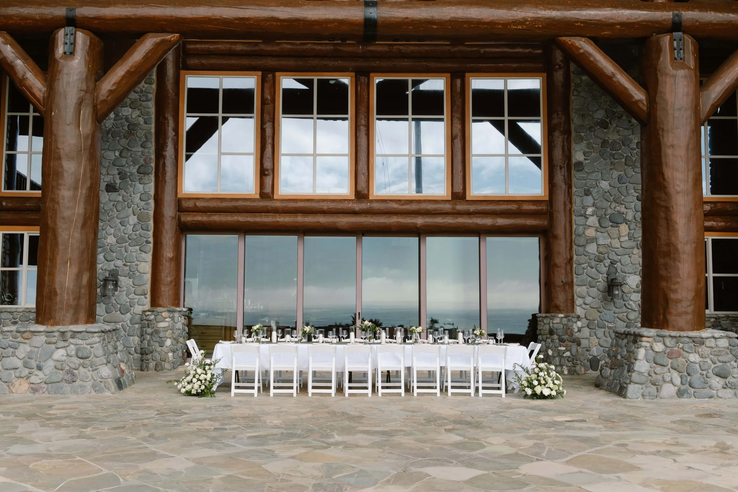 Long white reception table set beneath a stone and timber coastal lodge overlooking the ocean.
