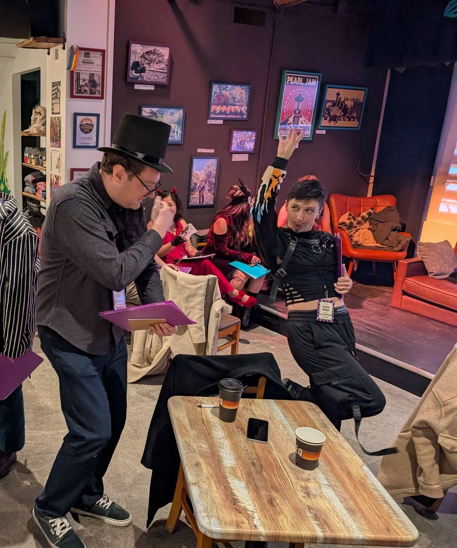 A player in costume dramatically raises an arm while another guest reads notes, creating a theatrical moment during the murder mystery.