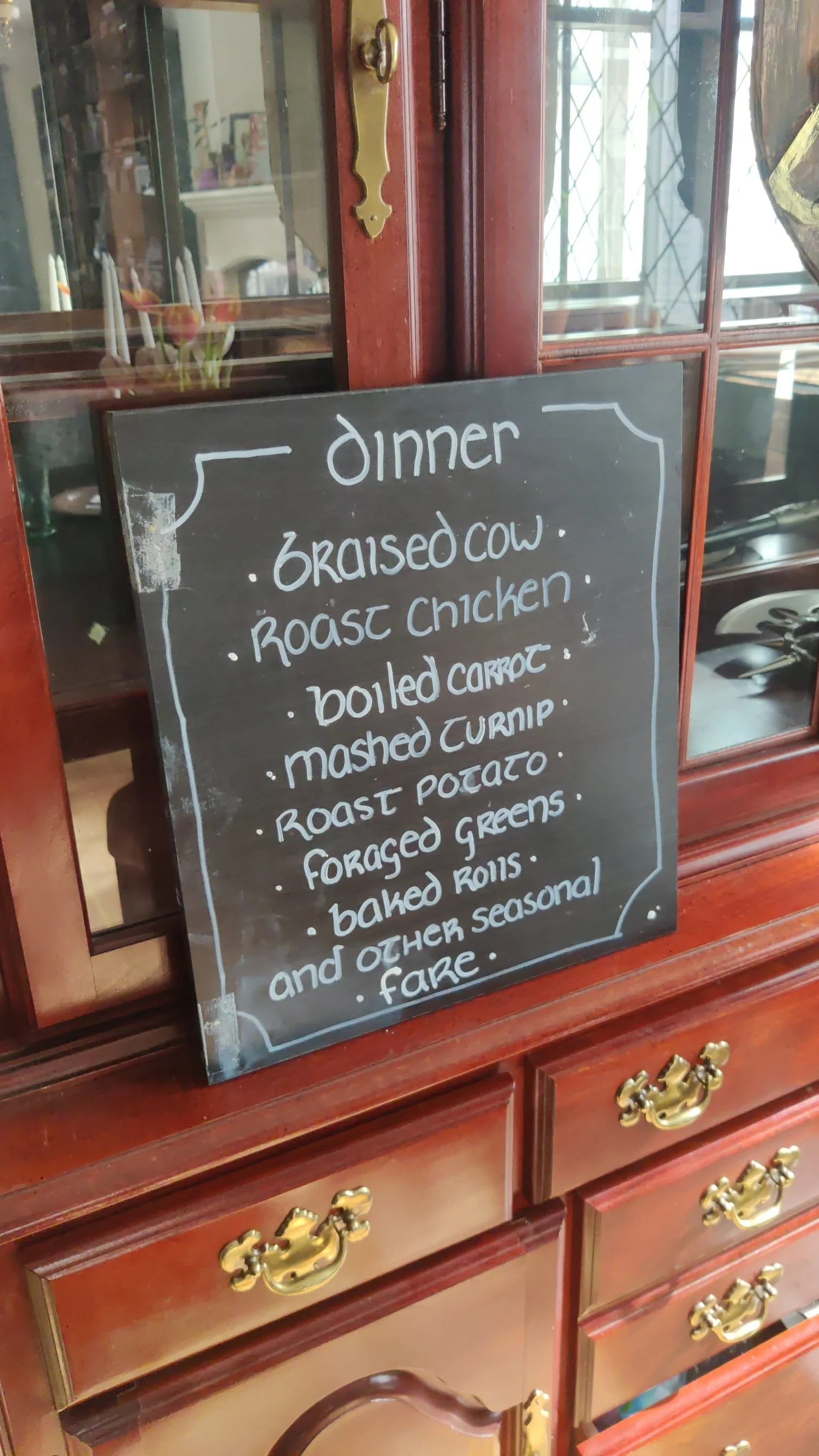 Fantasy murder mystery dinner menu featuring braised beef, roast chicken, root vegetables, and baked rolls displayed on a rustic chalkboard