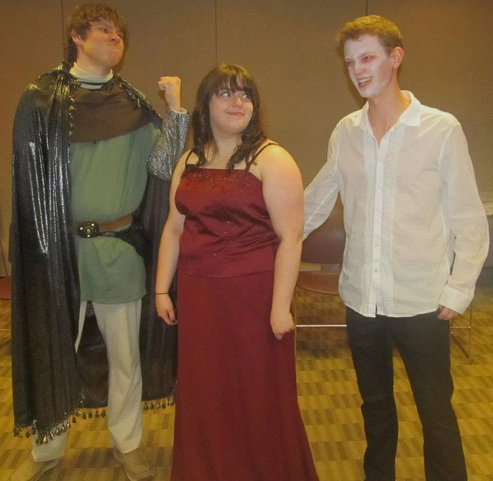 A group of three poses at a murder mystery game wearing medieval fantasy costumes. A knight shakes his fist as his partner looks lovingly at a vampire.