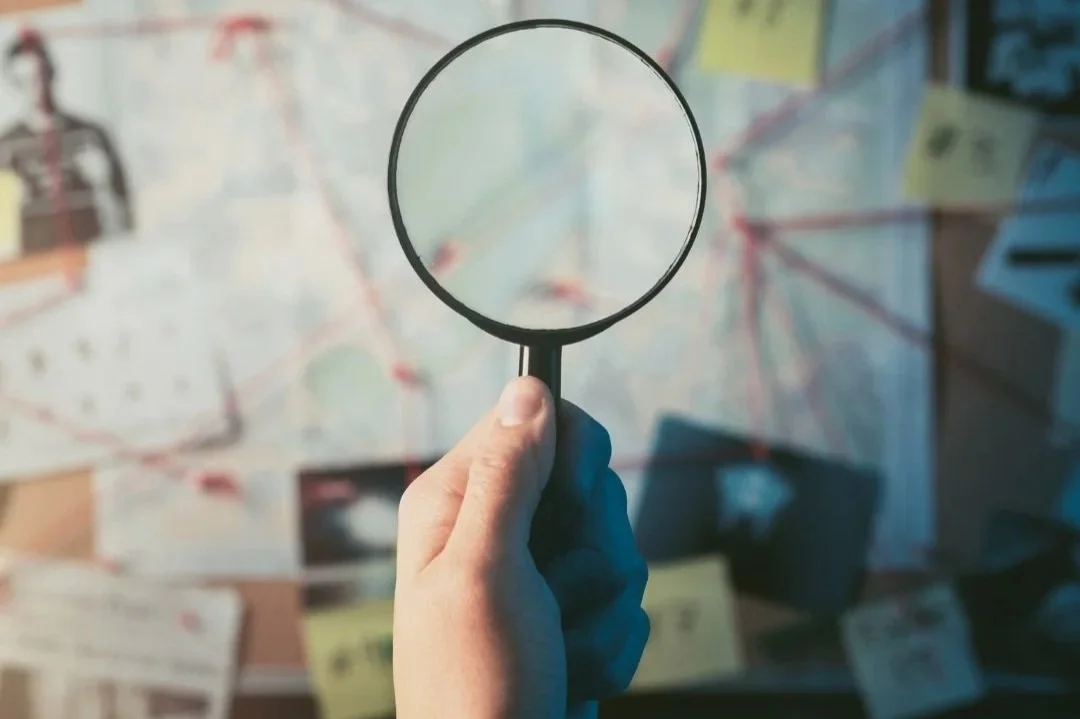 A hand holding a magnifying glass over a conspiracy mystery board with red string
