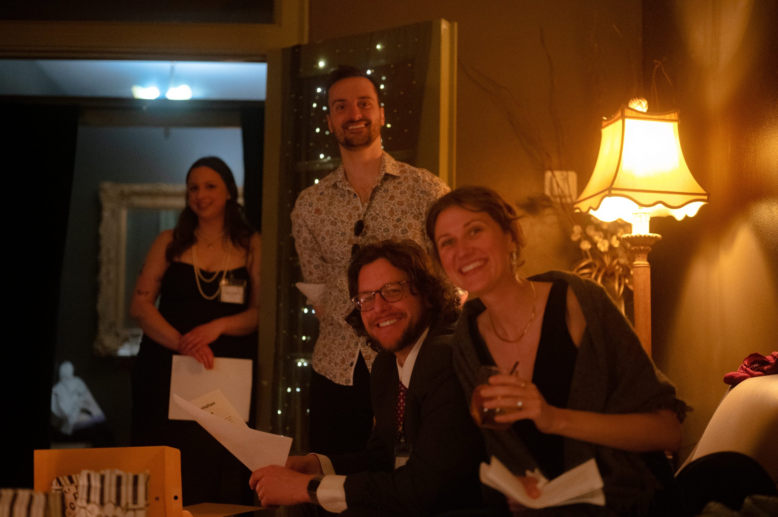 Friends laughing and holding game materials during a murder mystery party in a warmly lit living room, immersive Behind the Curtain gameplay moment