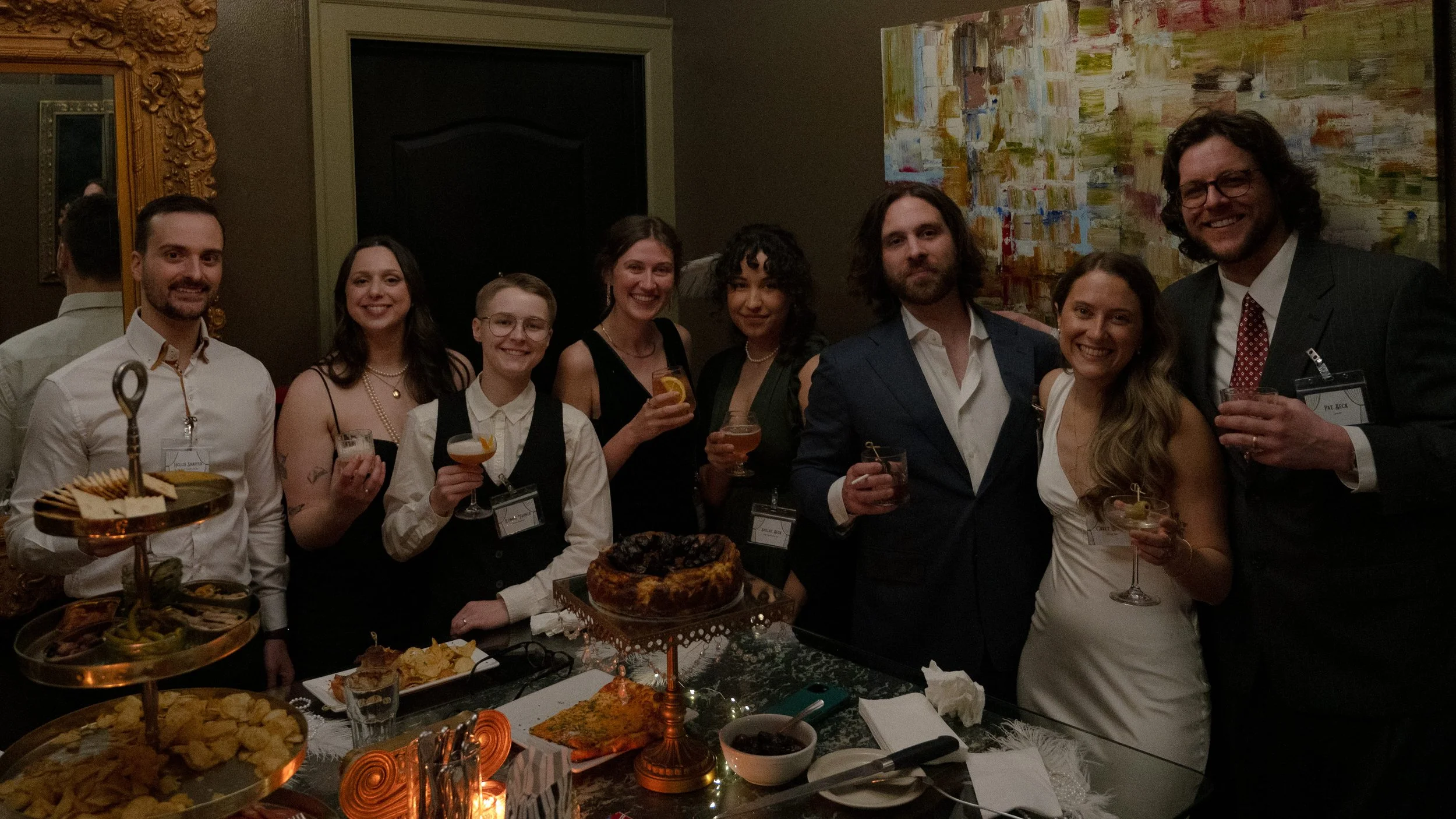 1950s Hollywood murder mystery party group dressed in vintage formal attire, holding cocktails around a themed party table with desserts and charcuterie, playing Behind the Curtain game
