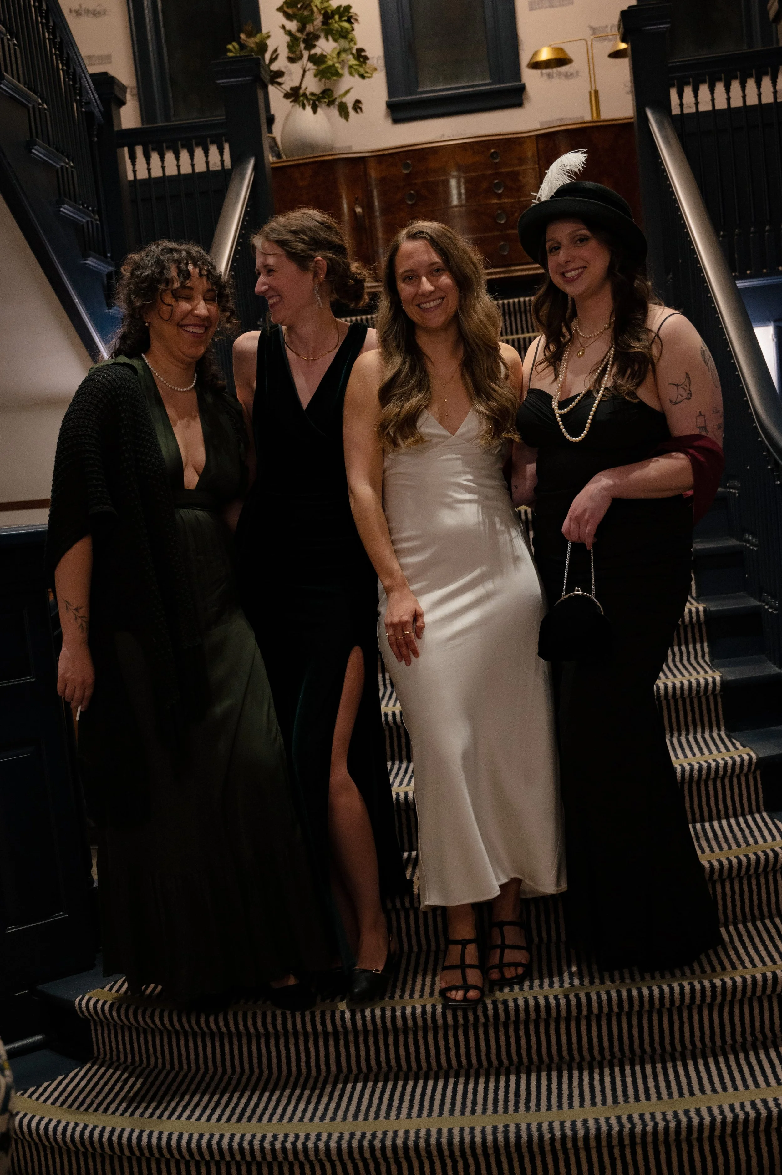 Group of women in elegant 1950s Hollywood dresses posing on staircase, vintage glam murder mystery party outfits for Behind the Curtain game night