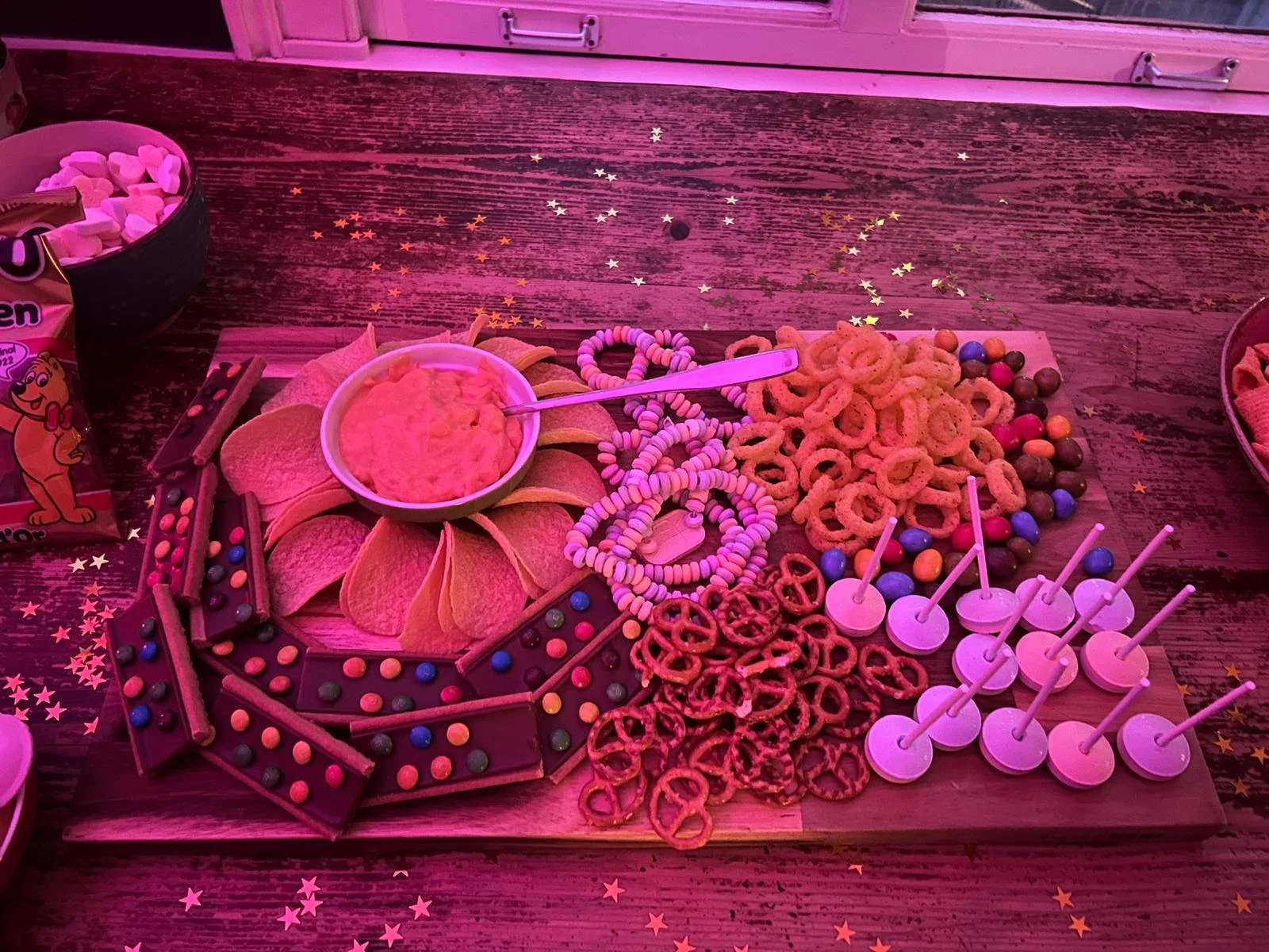 90s party food ideas with colorful snack board including chips and dip, pretzels, candy, chocolate bars, and cake pops for a themed murder mystery party