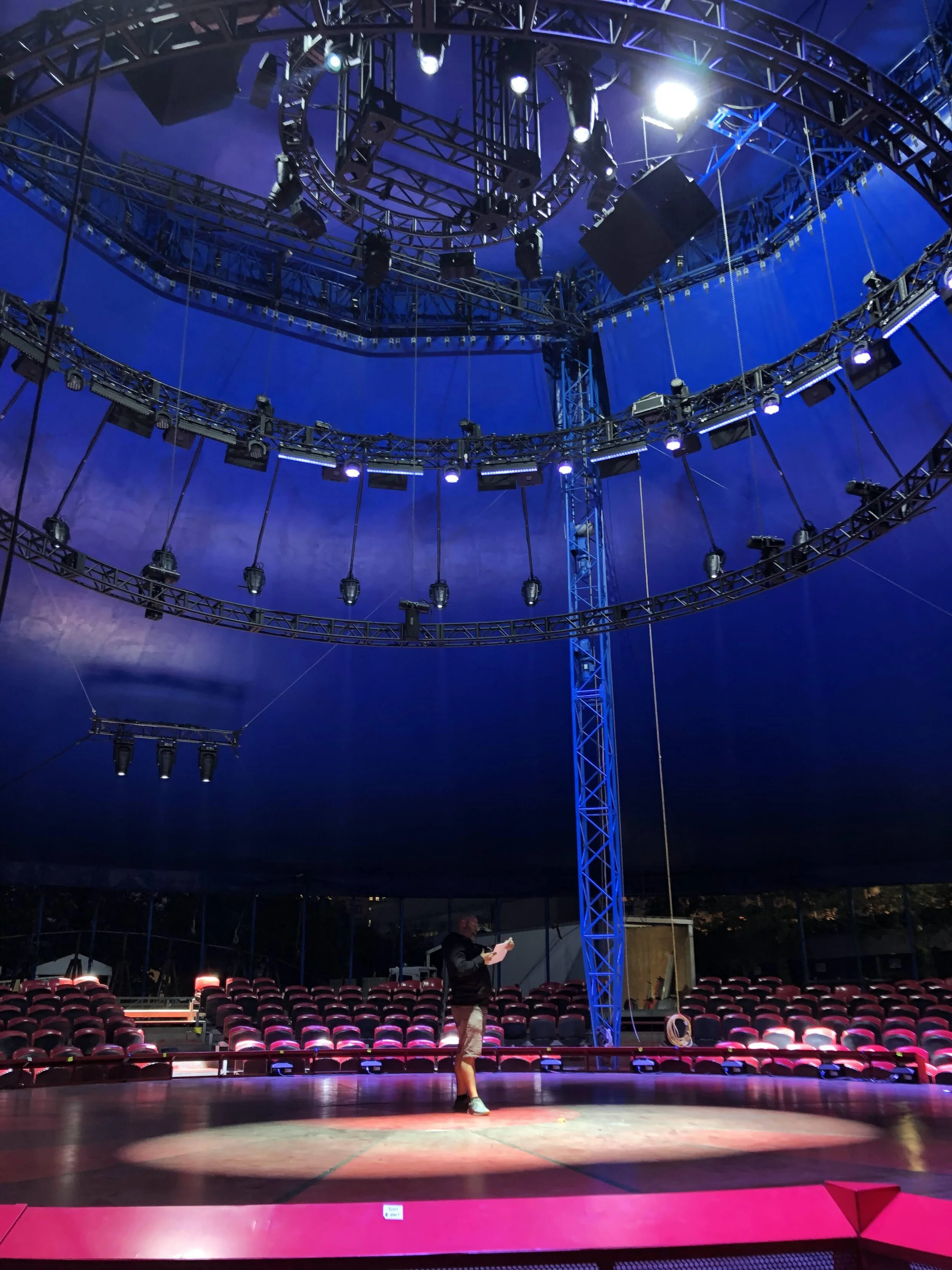 A man standing in the center of a circular stage under a large, blue canopy with stage lights overhead, and empty red seats surrounding the stage.