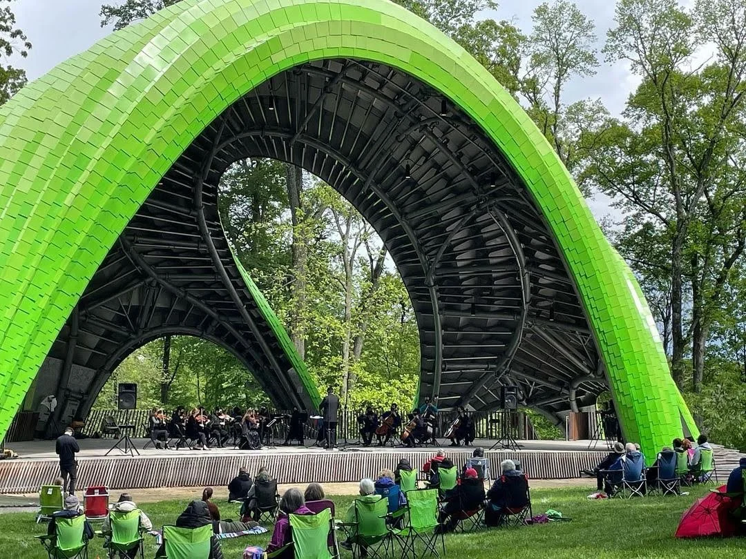 Outdoor concert in a park with a green, arch-shaped stage and a small orchestra performing, audience seated on lawn chairs.