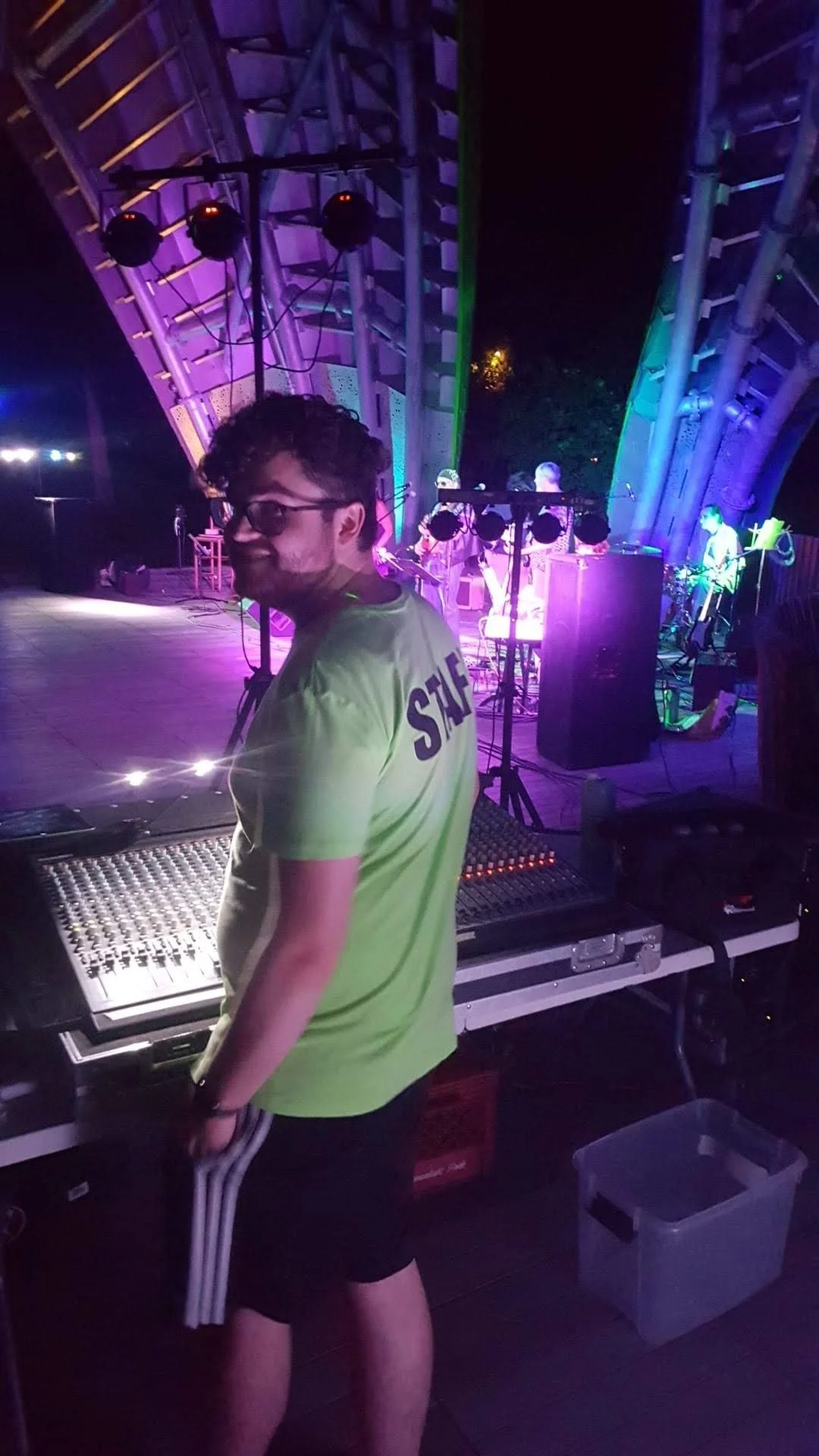 A young man wearing glasses and a neon yellow staff shirt stands at a mixing console in a dimly lit stage setting with colorful lights, with a band performing in the background.