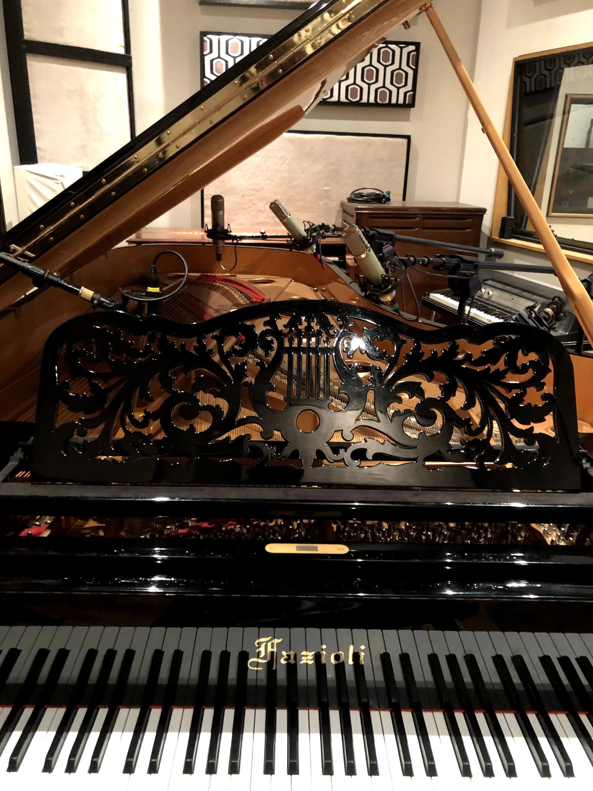 Inside a music recording studio, there is a grand piano with a decorative music stand, microphone setup, and framed acoustic panels on the wall.