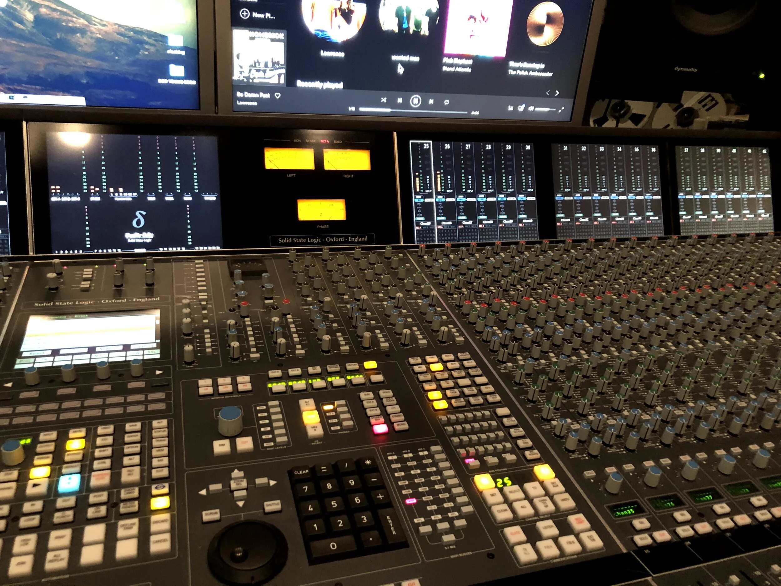 A professional sound mixing console with numerous faders, knobs, and buttons, with multiple monitors displaying audio levels and media interfaces in the background.
