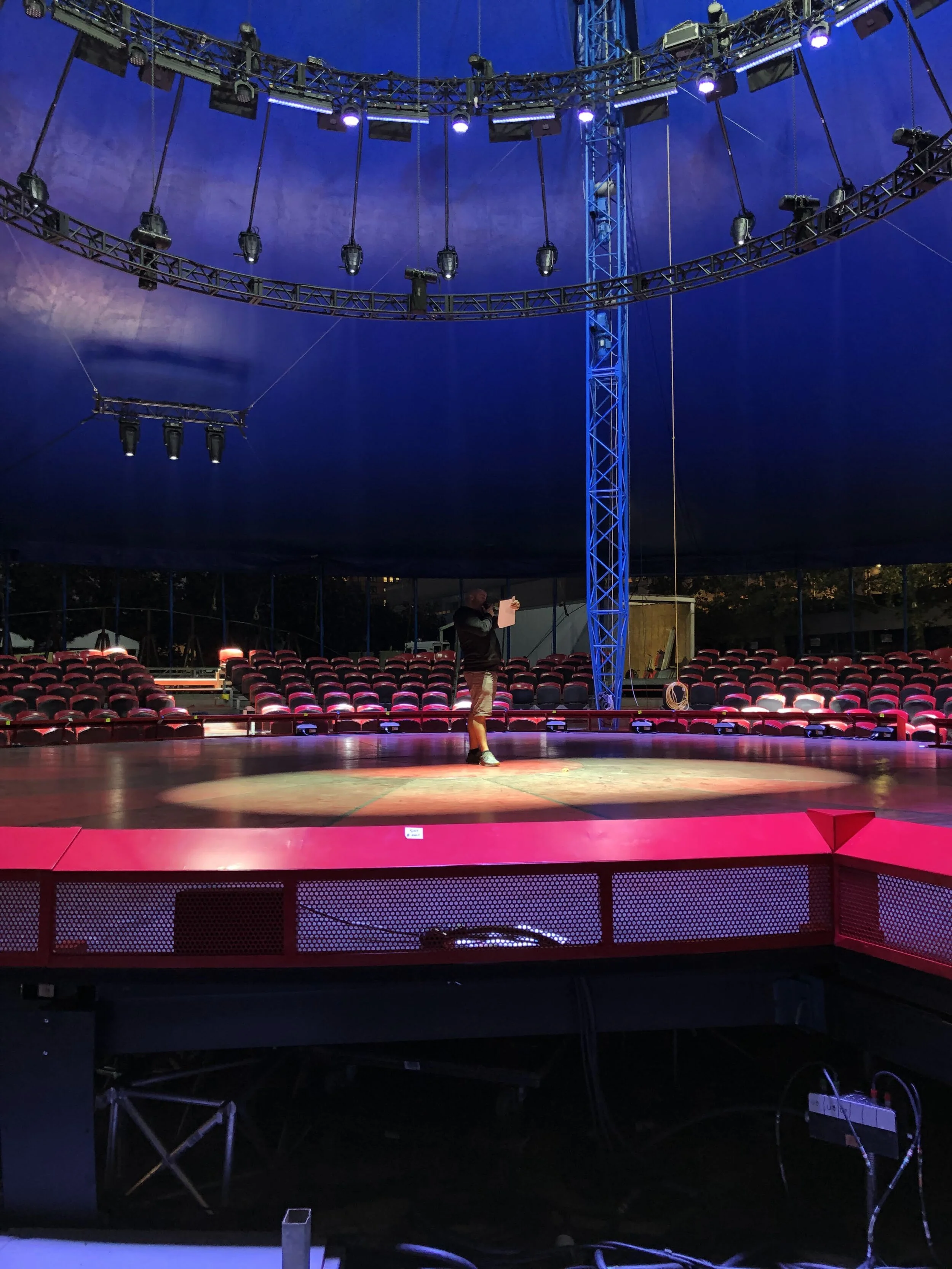 Interior of a circus tent with a raised stage, surrounded by red seating, lit with purple and pink lights, and a person standing on stage holding a paper.