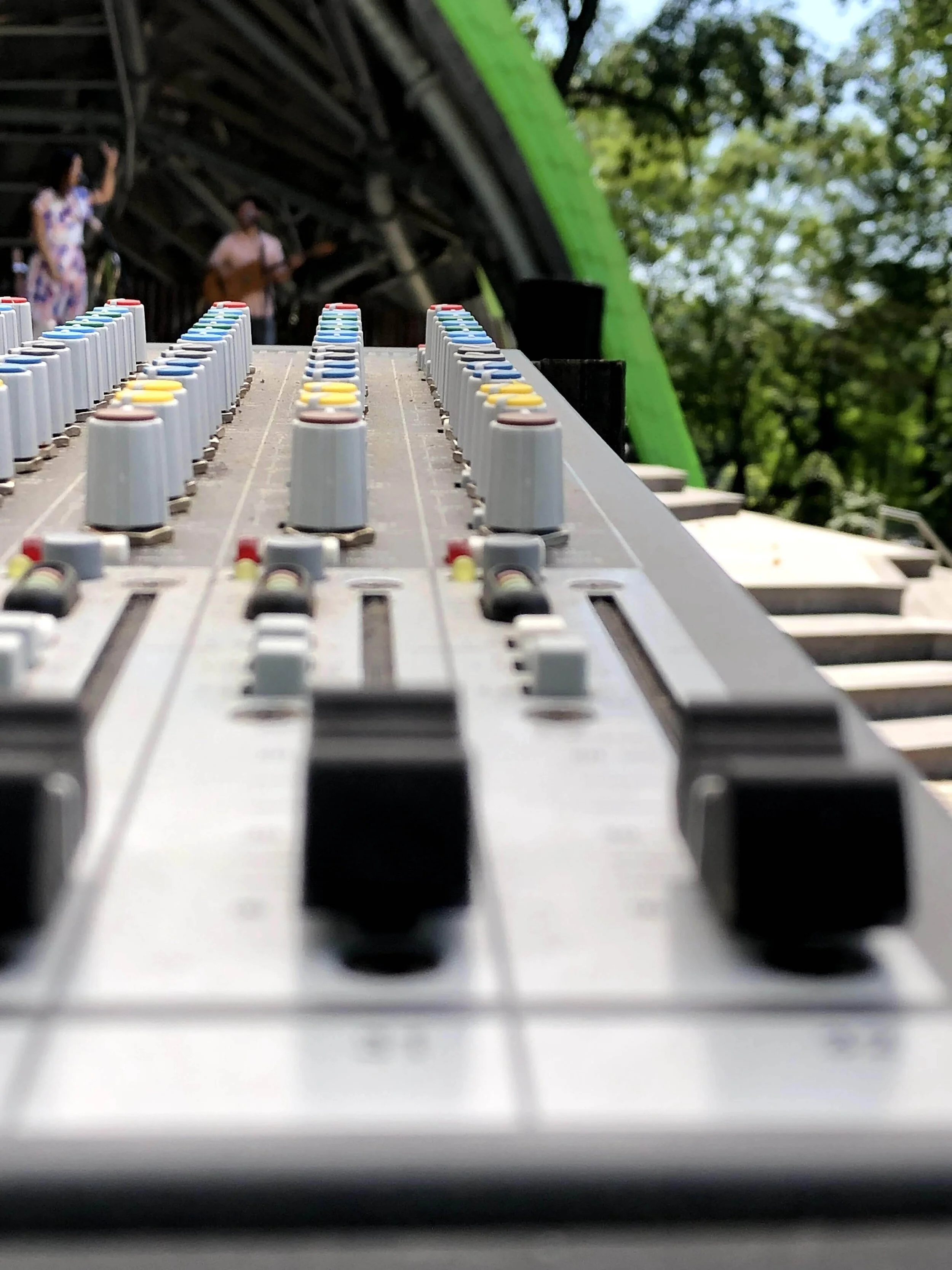 Close-up view of a sound mixing console with blurred band performing on an outdoor stage in the background.