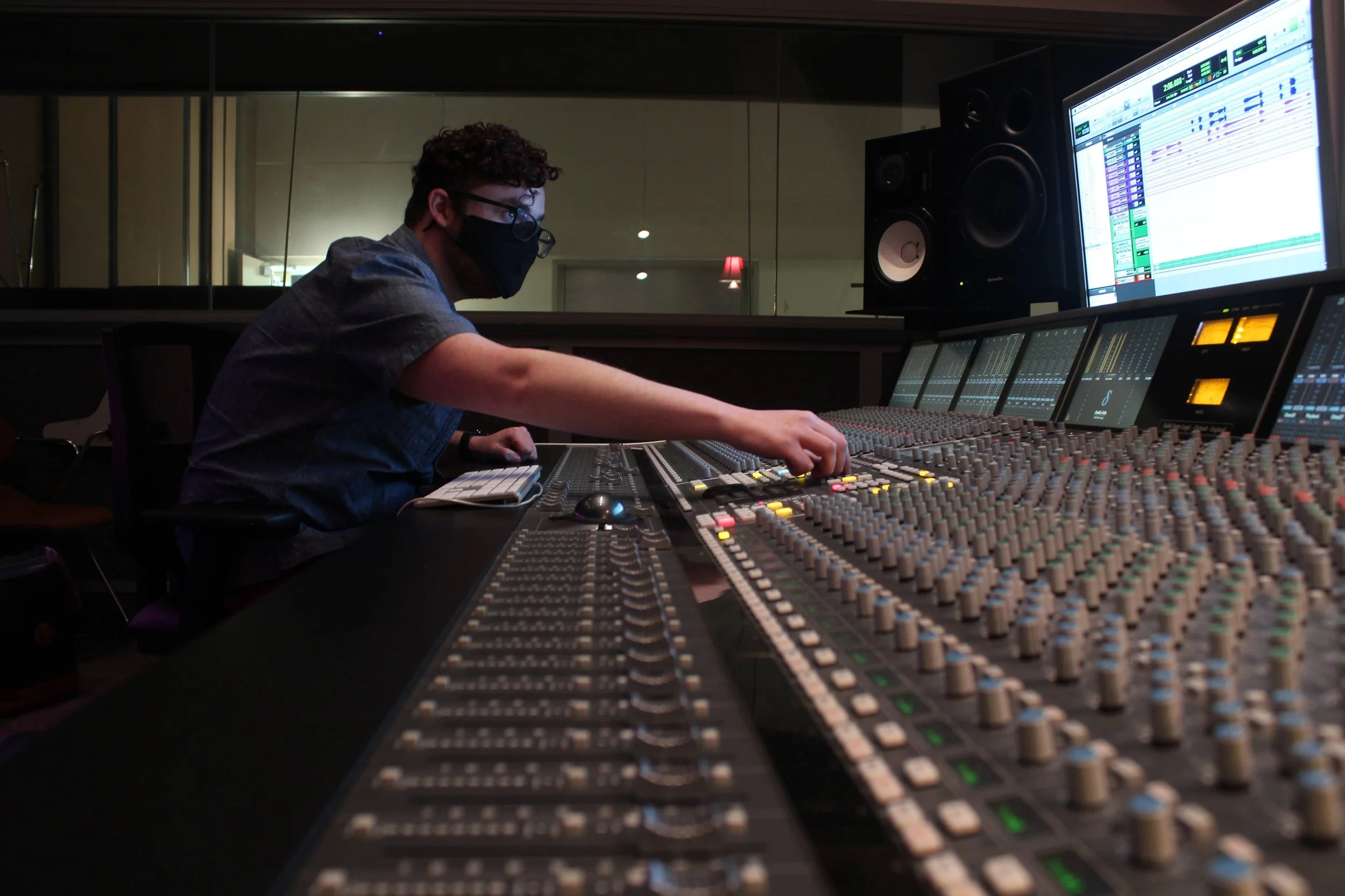A person wearing a black face mask and glasses working at a large audio mixing console with multiple screens and audio equipment.