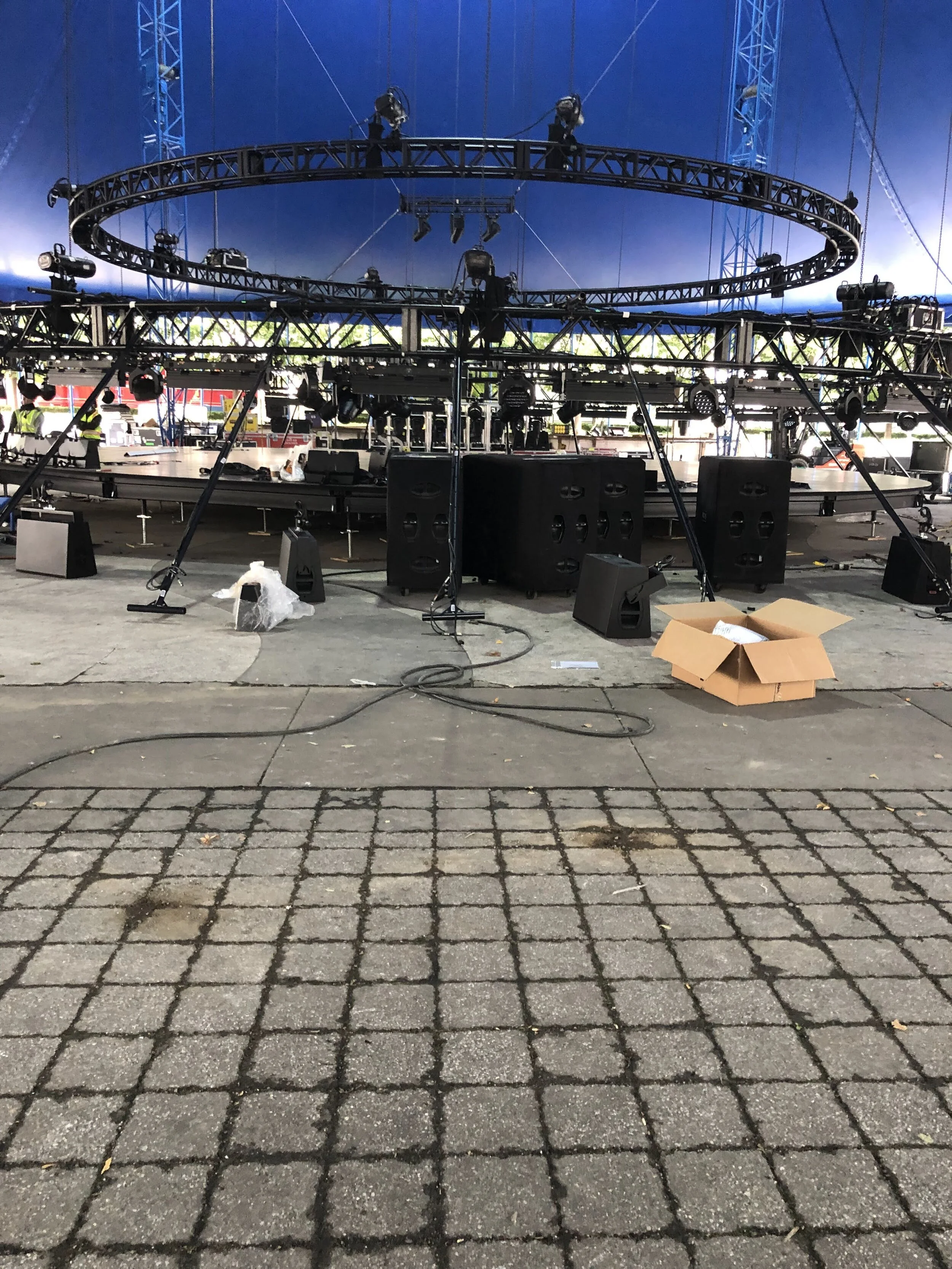 Stage setup with lighting and sound equipment under a blue tent, with a cardboard box on the ground and cobblestone pavement in the foreground.