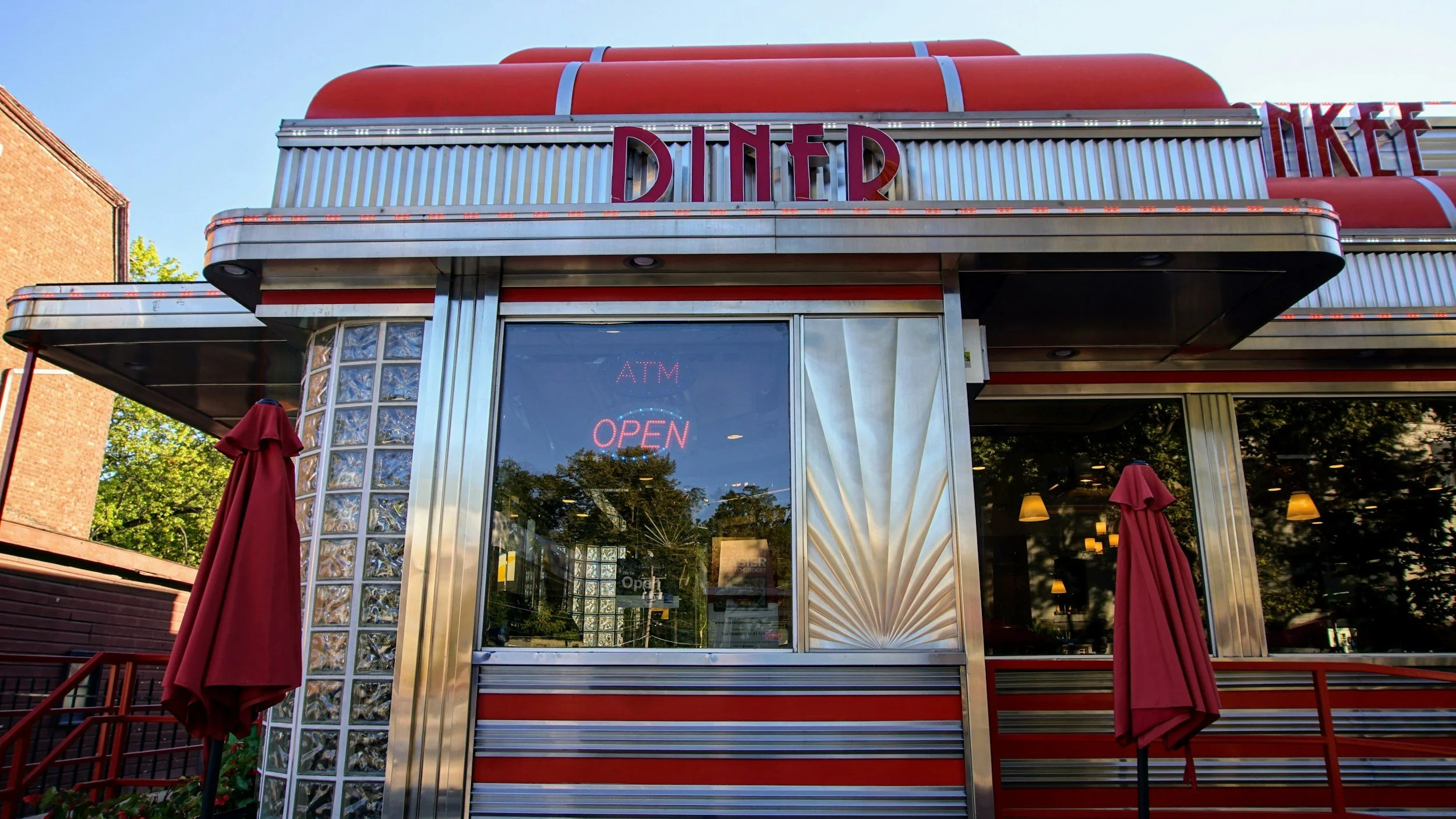 Exterior of a vintage diner with red and silver design, illuminated sign reading 'ATM OPEN,' and red umbrellas outside.