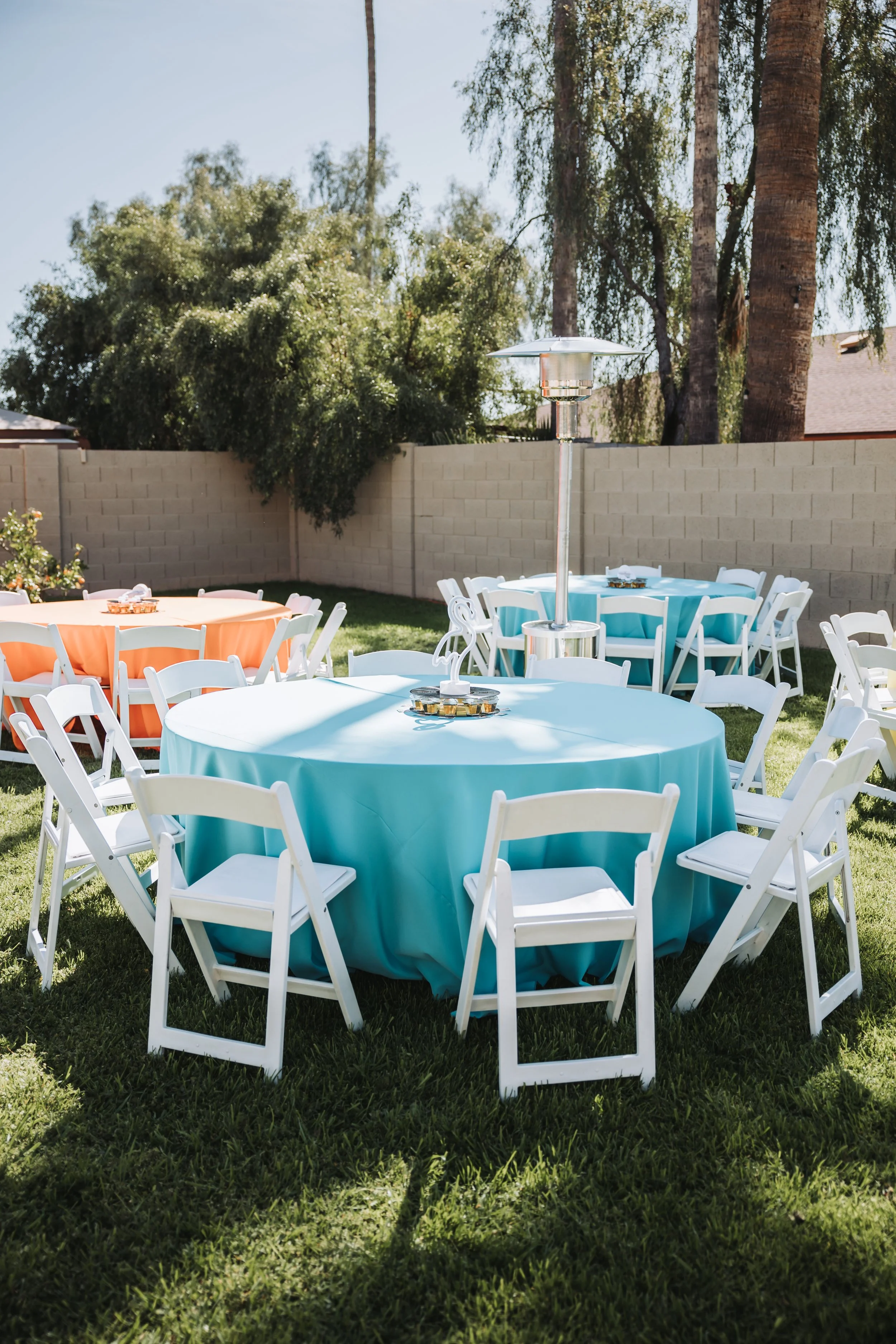 Round tables covered with teal and orange tablecloths and white folding chairs set up on a lawn, with a patio heater in the center, in a backyard with a brick wall and tall trees.