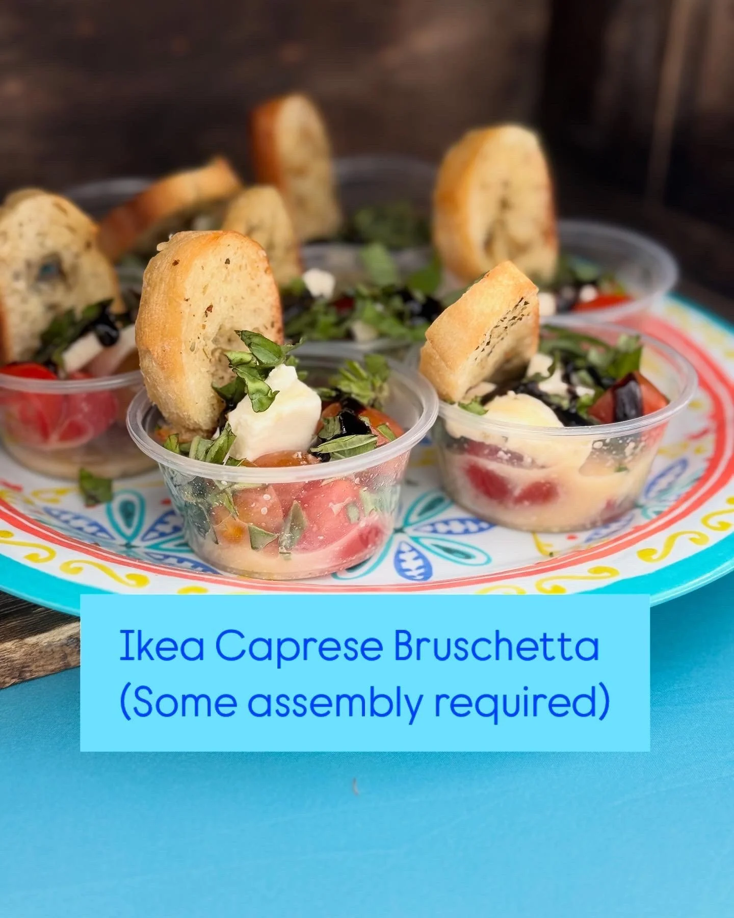 Small clear cups containing caprese salad with sliced tomatoes, mozzarella, basil, black olives, and toasted baguette slices, arranged on a colorful plate, labeled 'Ikea Caprese Bruschetta (Some assembly required)'.