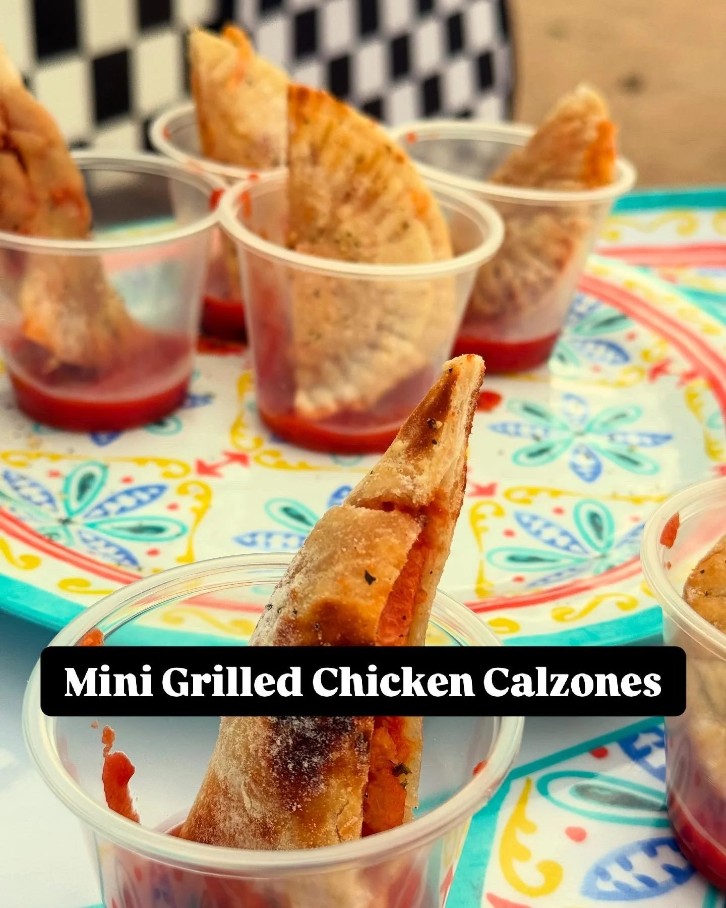 Mini grilled chicken calzones in clear cups on a colorful patterned tray with a black and white checkered backdrop.