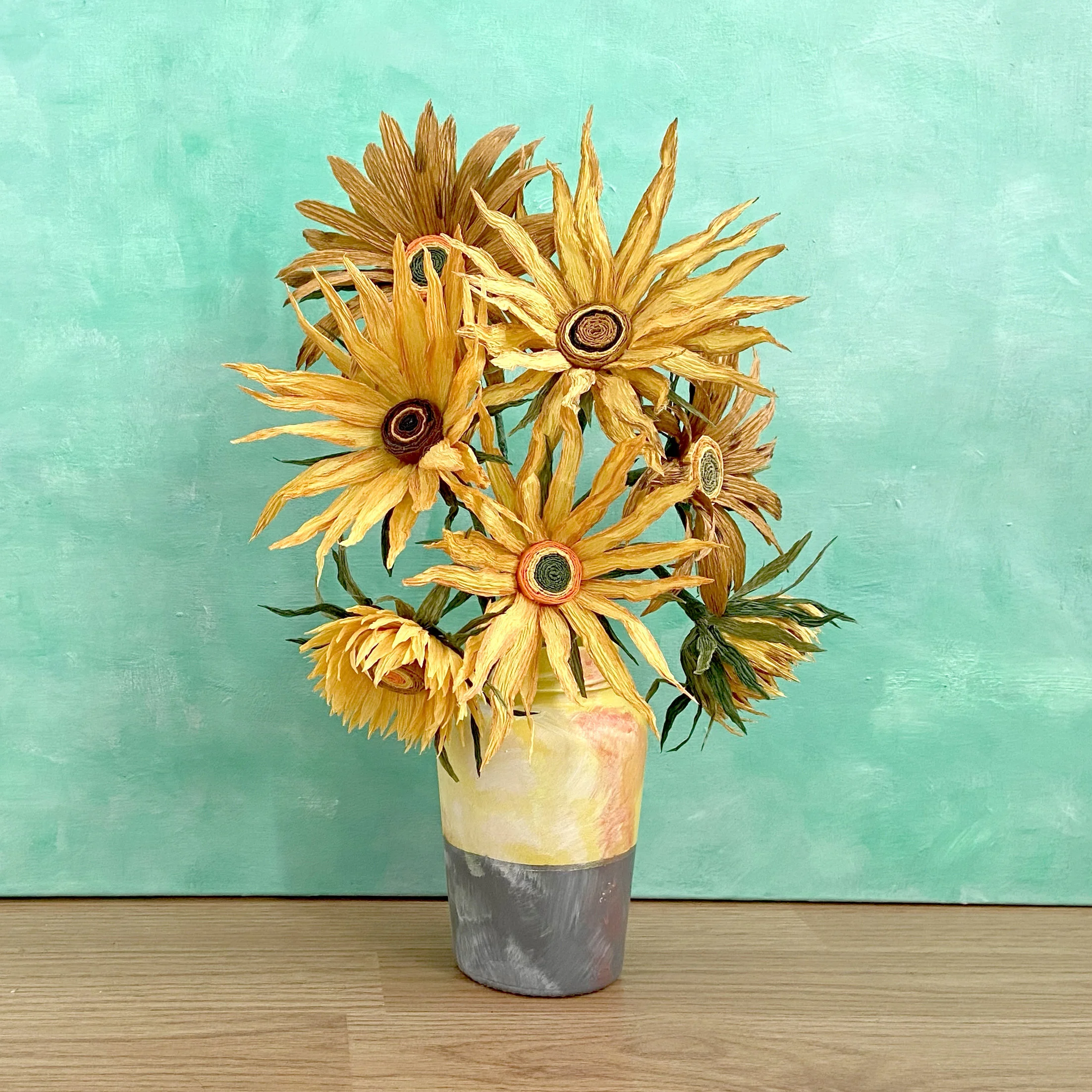 Artificial sunflower bouquet in a multicolored vase on a wooden surface against a teal background.