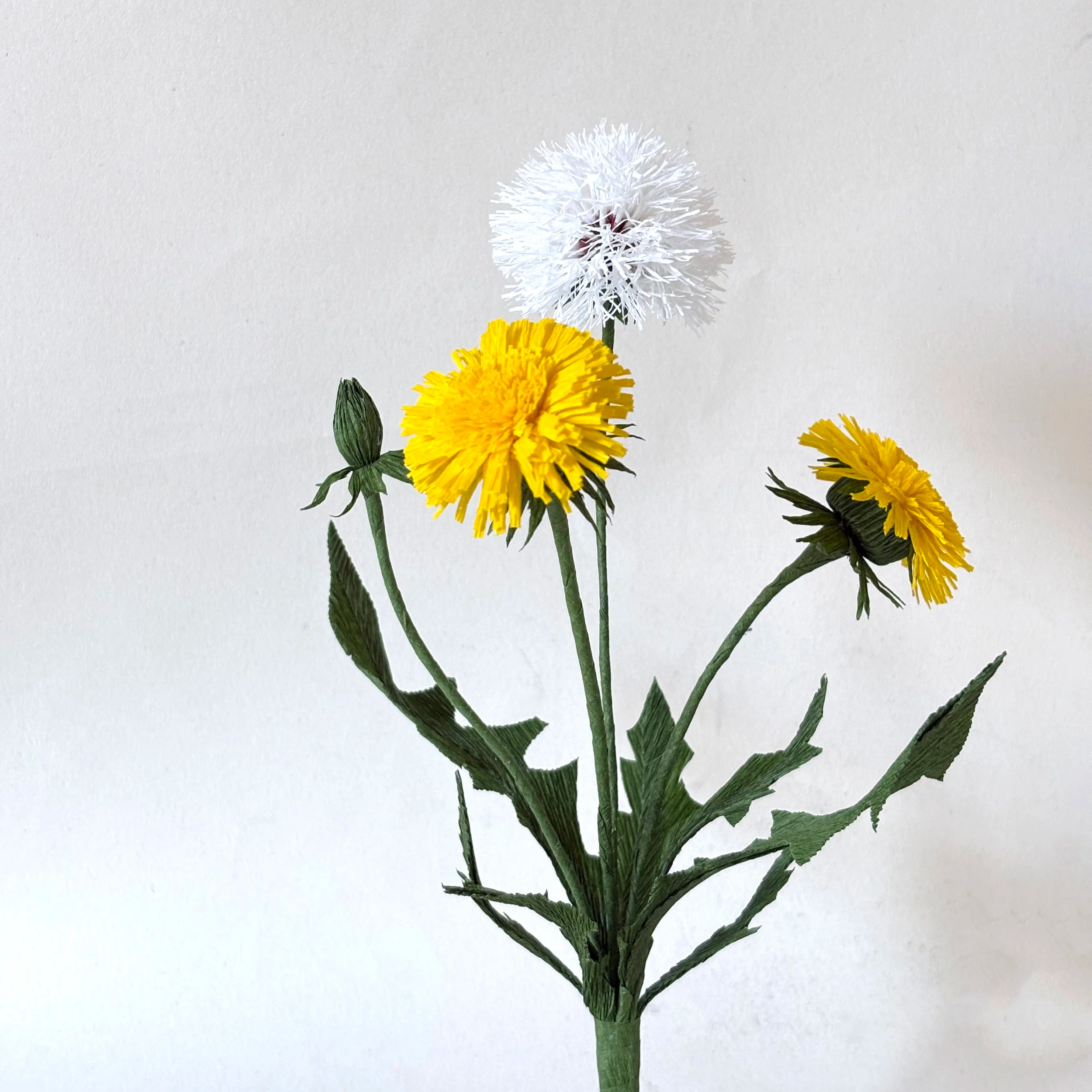 A bouquet of yellow dandelion flowers with green stems and leaves, featuring a white artificial flower in the middle, set against a plain white background.