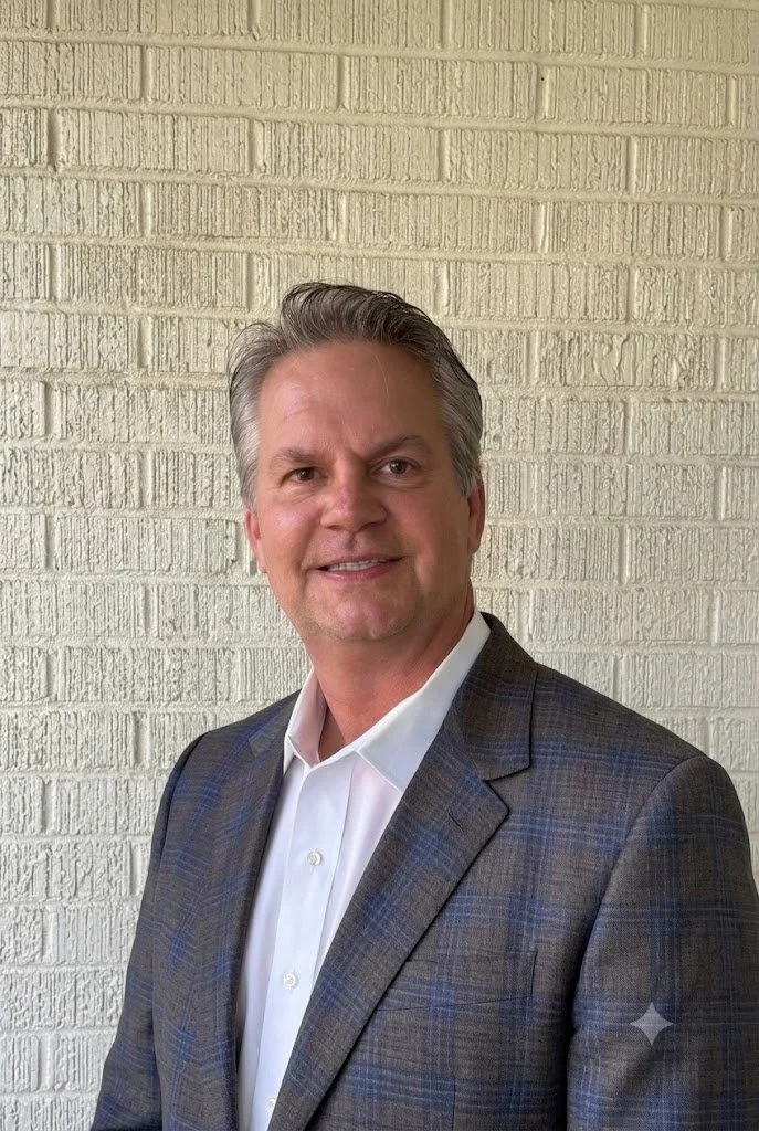 A middle-aged man with gray hair, wearing a white dress shirt and a dark plaid suit jacket, standing in front of a beige brick wall.