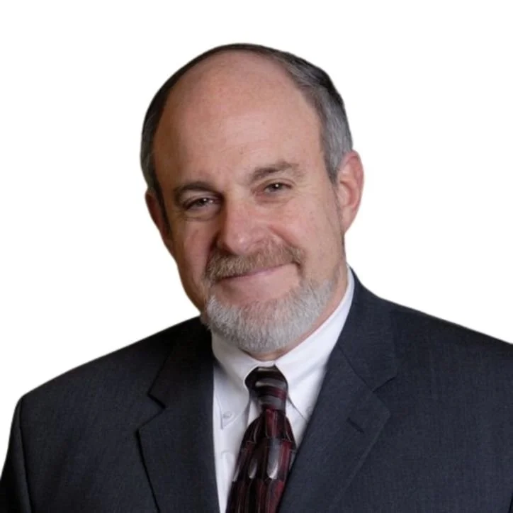 Headshot of an older man with a beard, wearing a dark suit, white shirt, and patterned tie, smiling against a plain white background.