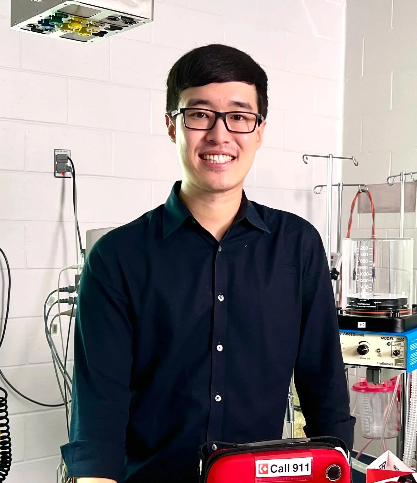 A man with black hair and glasses wearing a dark shirt smiling in a medical setting. Medical equipment including a defibrillator with a sign that says 'Call 911' is visible behind him.