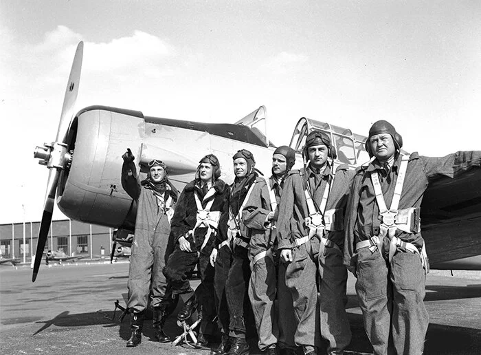 A posed photograph of American student pilots of the British Commonwealth Air Training Plan at No. 2 SFTS Uplands, Course 25 in the spring of 1941. This was in the weeks leading up to the filming at Uplands of major sequences of Captains of the Clouds, Warner Bros.’ largest production to date. Starring James Cagney, the producers of the film looked to a premier in New York early in 1942. At left is Leading Aircraftman John Magee pointing to some lofty goal with posed determination and perhaps a bit of embarrassment. Perhaps this promotional shot was destined to inspire Americans to step up and get behind the Allies in the expedition of the war. While the film was in post-production, the Japanese attack on Pearl Harbor eliminated any need for the film to encourage the Yanks. Captains of the Clouds opened in February of 1942 but America hardly noticed, preoccupied as it was with war preparations. The pilots are (L to R): Aircraftmen John G. Magee of Washington (J5824); Arthur C. Young of Cleveland, Ohio; Claiborne Frank Gallicher of Tulsa, Oklahoma; Curtis Gilman Johnston of Chicago, Illinois; Arthur Bernard Cleaveland of Springfield, Illinois; and Ober Nathaniel Leatherman of Lima, Ohio. Photo: DND