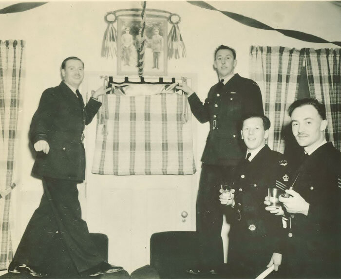 Squadron Leader W.C. Connell, commanding officer, (Left) stands on a mess chair ready to lower a plaid curtain to reveal to staff photographers, the new 135 Squadron fighting bull dog emblem at their new home in Patricia Bay, Vancouver Island in 194…