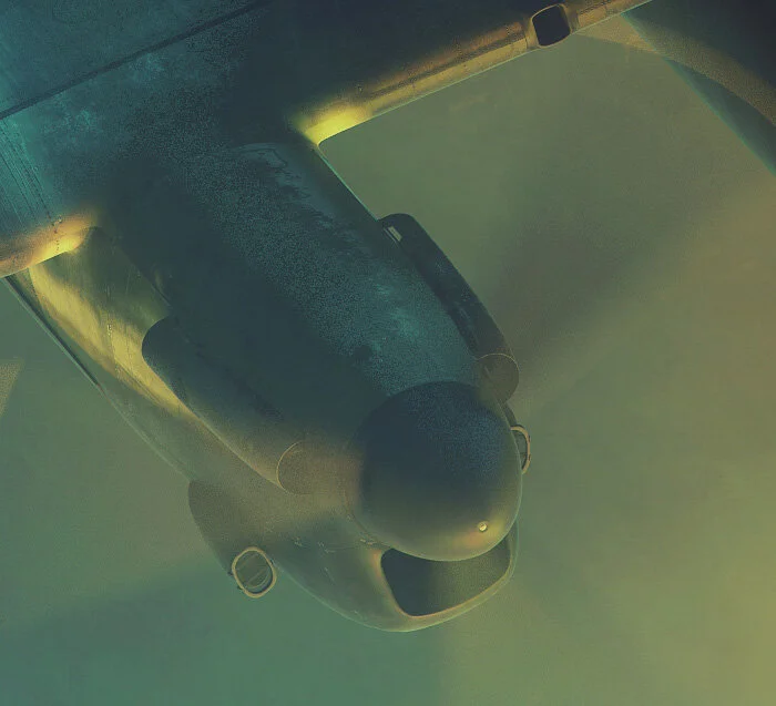 The blue light of searchlights and the yellow fires below combine in an eerie green light, as we gaze downward, past the Number 2 engine of AS-J2. The beams of searchlights and their effects on smoke and cloud present many problems to a digital artist looking to accurately portray a scene over a burning city. Forkasiewicz is careful to allow certain details, such as searchlight beams and other aircraft in the formation, to remain secondary to the central image. Detail from Toward the Inferno by Piotr Forkasiewicz