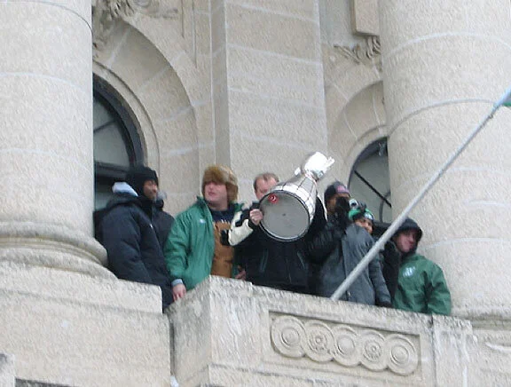 Two days later, the Saskatchewan Roughriders make a Papal-style appearance from the central balcony of the Saskatchewan Legislative Assembly building in Regina. With happily frozen Rider fans cheering wildly, team members raise the Grey Cup so that fans can see the famous sticker. Photo by computer_saskboy at Flickr.com