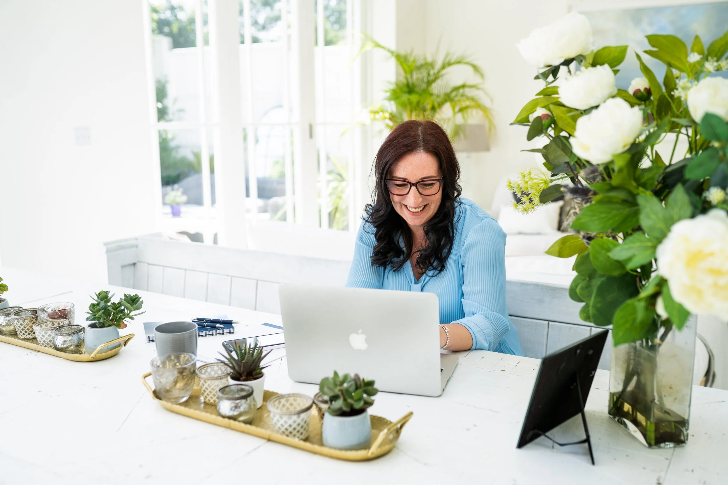 Team member working on a laptop at a desk in a bright room, surrounded by plants and decorative items.