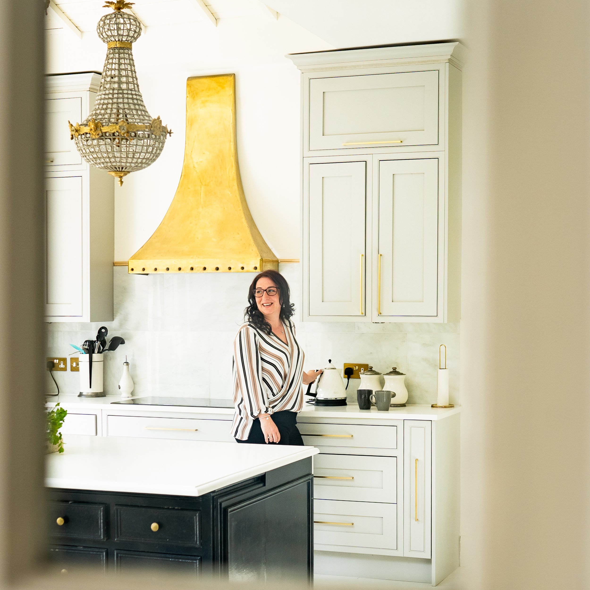 Woman wearing glasses standing in a bright, modern kitchen, smiling while preparing drinks beside white cabinetry and a gold cooker hood.