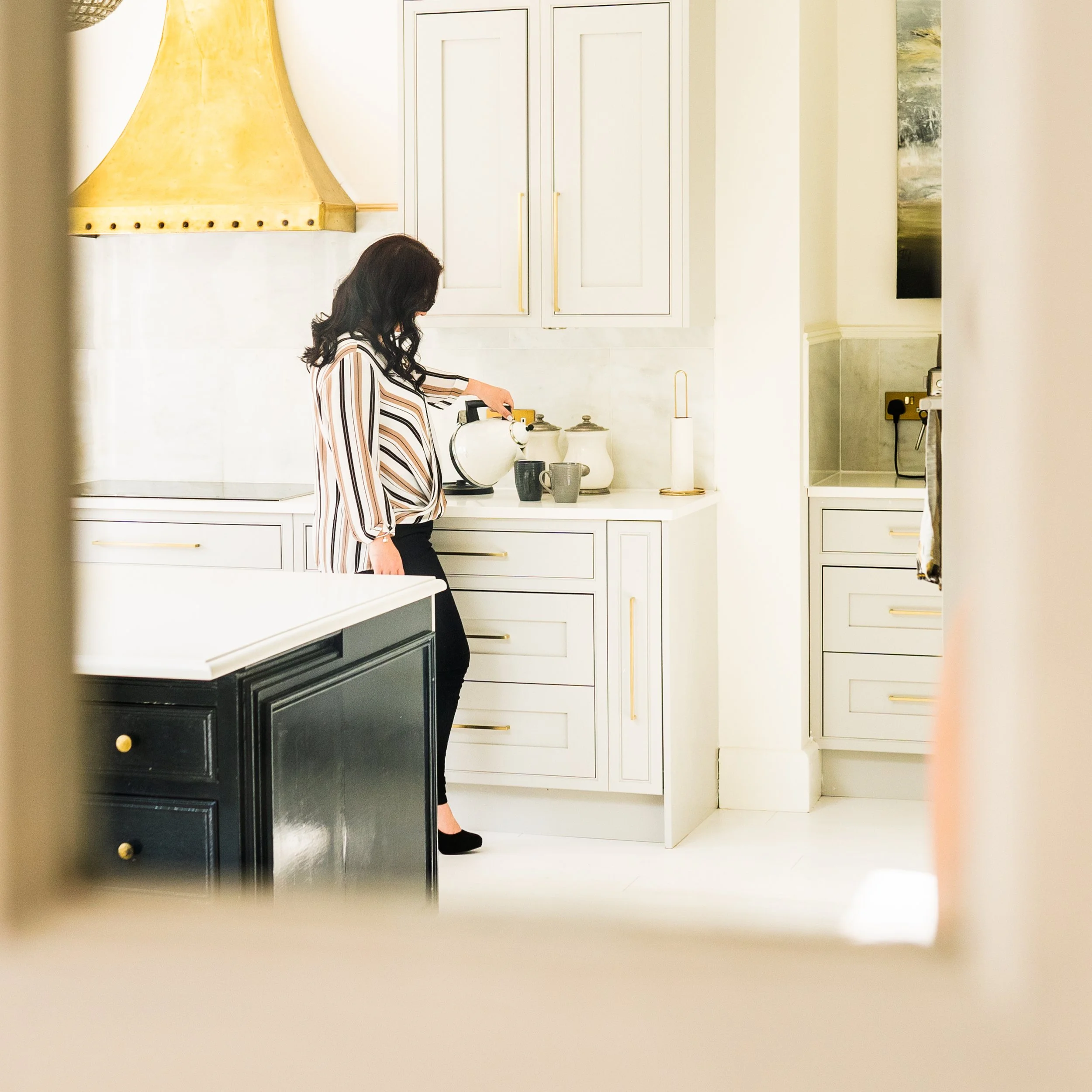 Businesswoman preparing tea in a stylish home kitchen with white cabinets and gold accents.