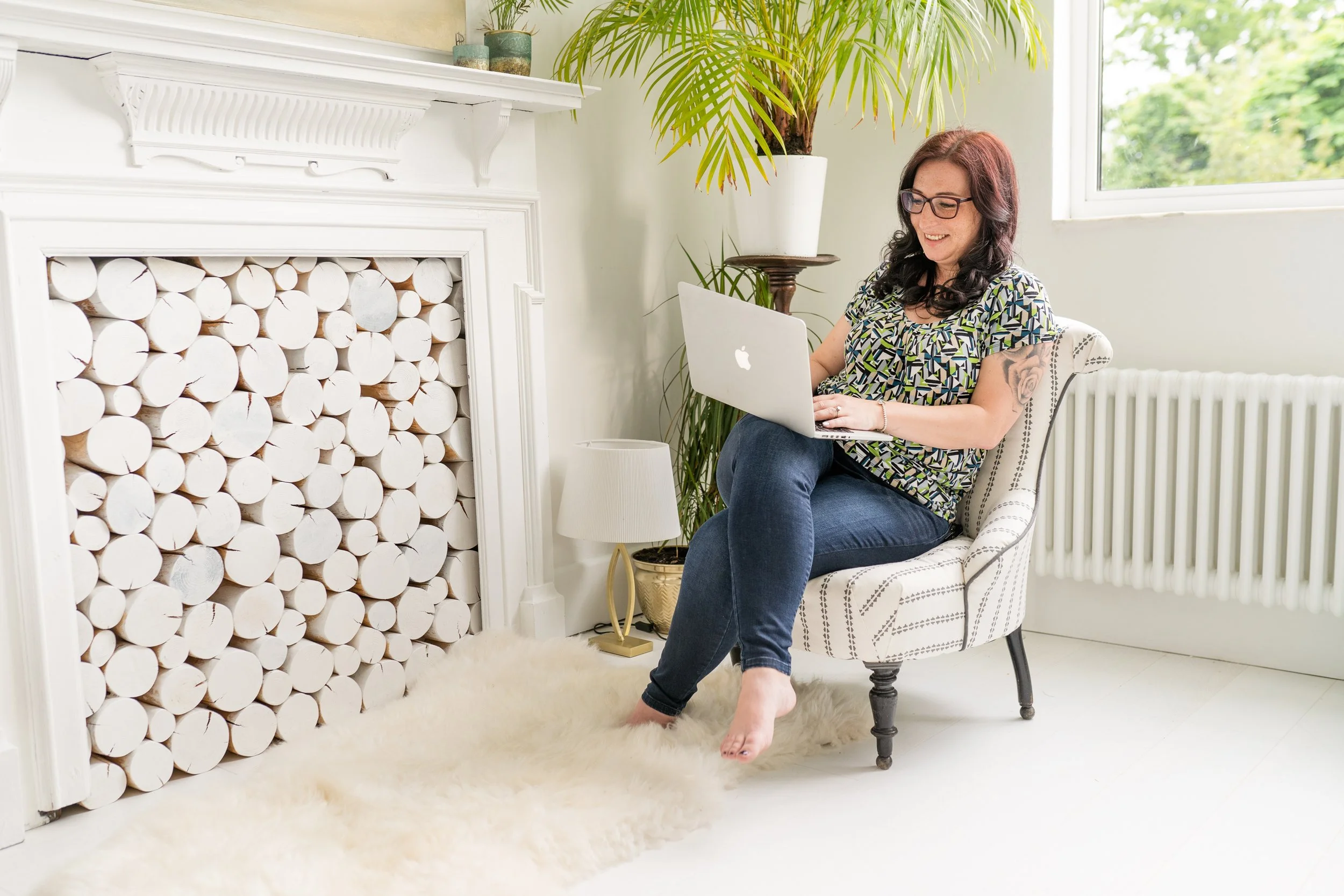 Woman sitting in an armchair using a laptop beside a white fireplace and large houseplant in a bright room.