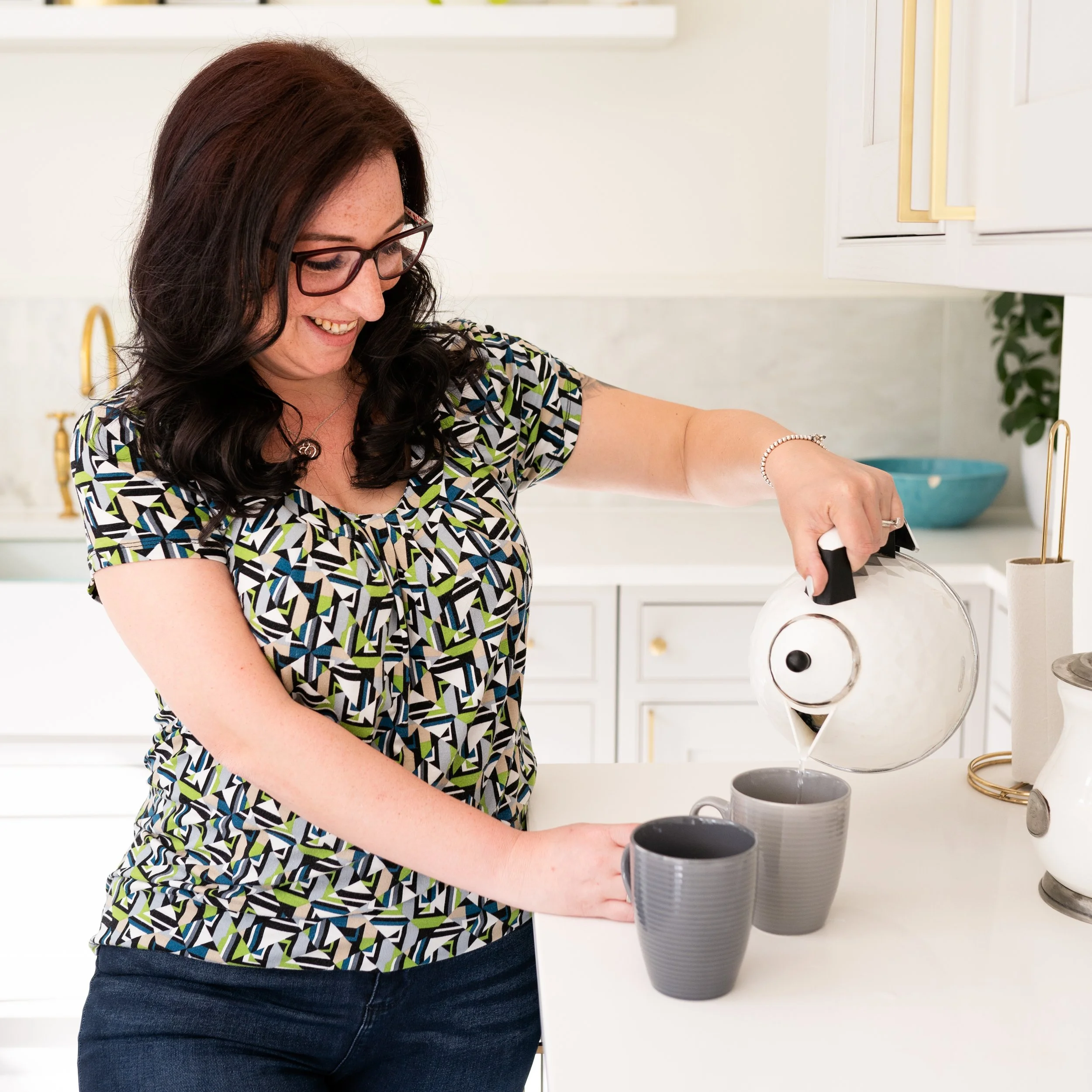 Woman wearing glasses smiling while pouring hot water from a kettle into two mugs in a bright kitchen.