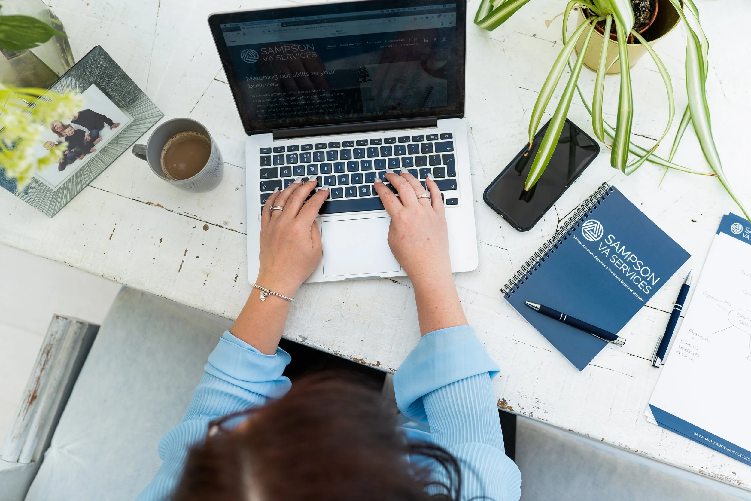 Overhead view of a woman working on a laptop at a white desk with notebook, phone and coffee.