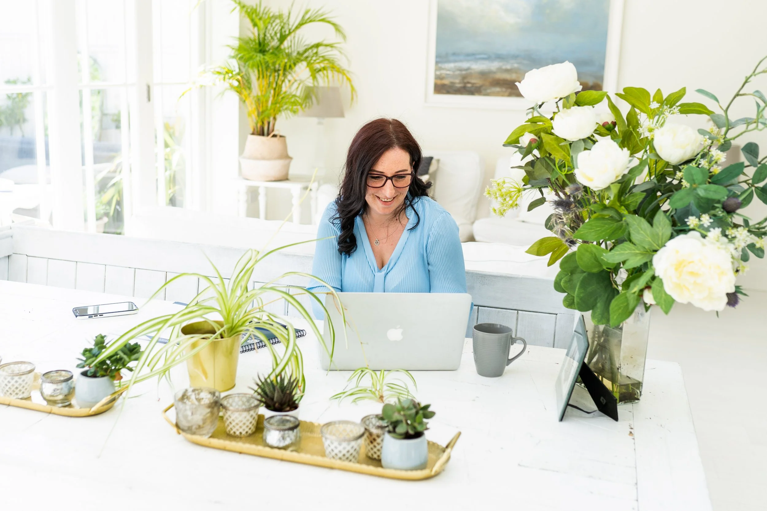Woman wearing glasses working on a laptop at a white table in a bright room filled with plants and flowers.