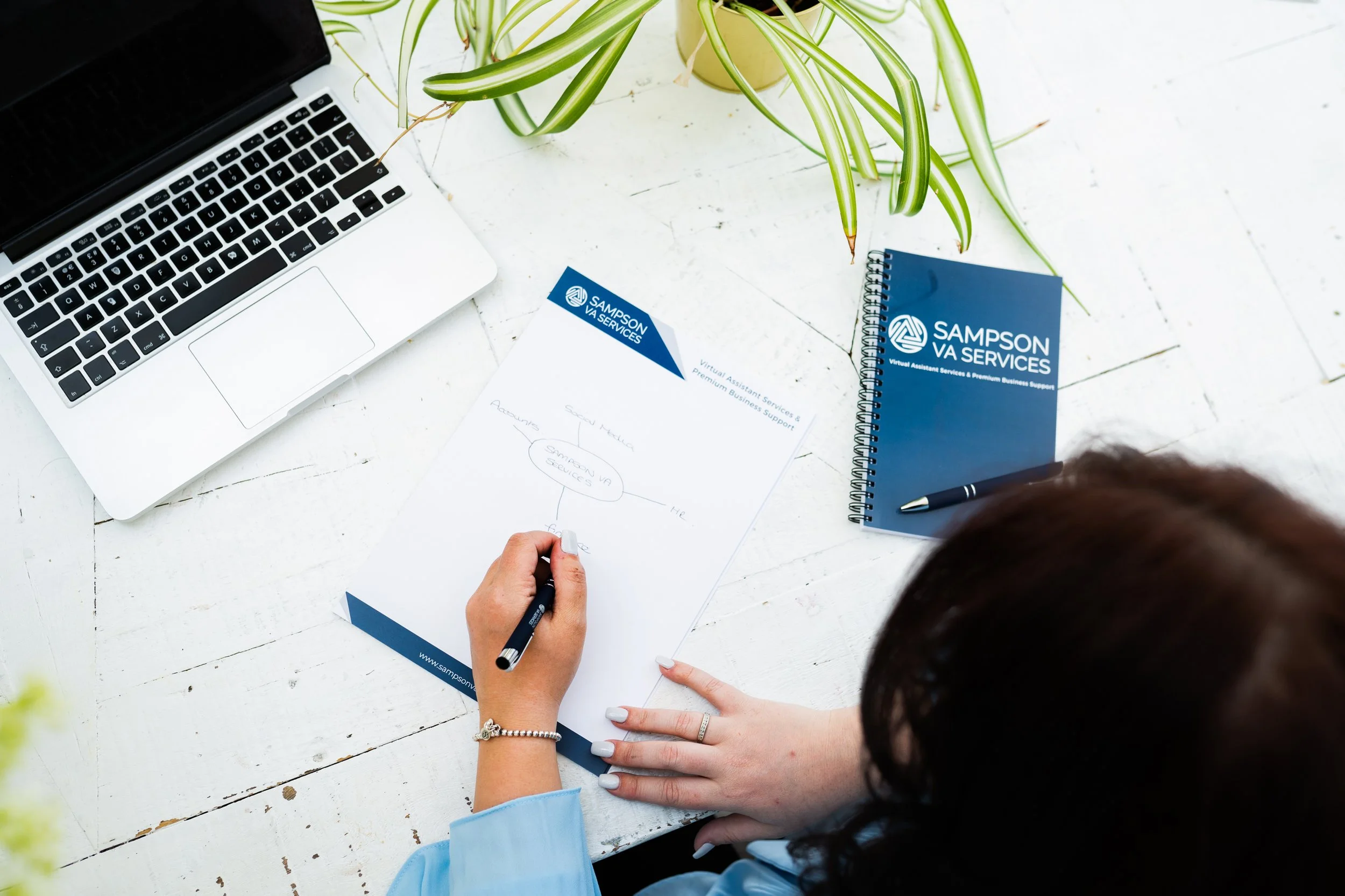 Overhead view of a team member writing notes on Sampson VA Services branded paperwork beside a laptop and notebook on a desk.
