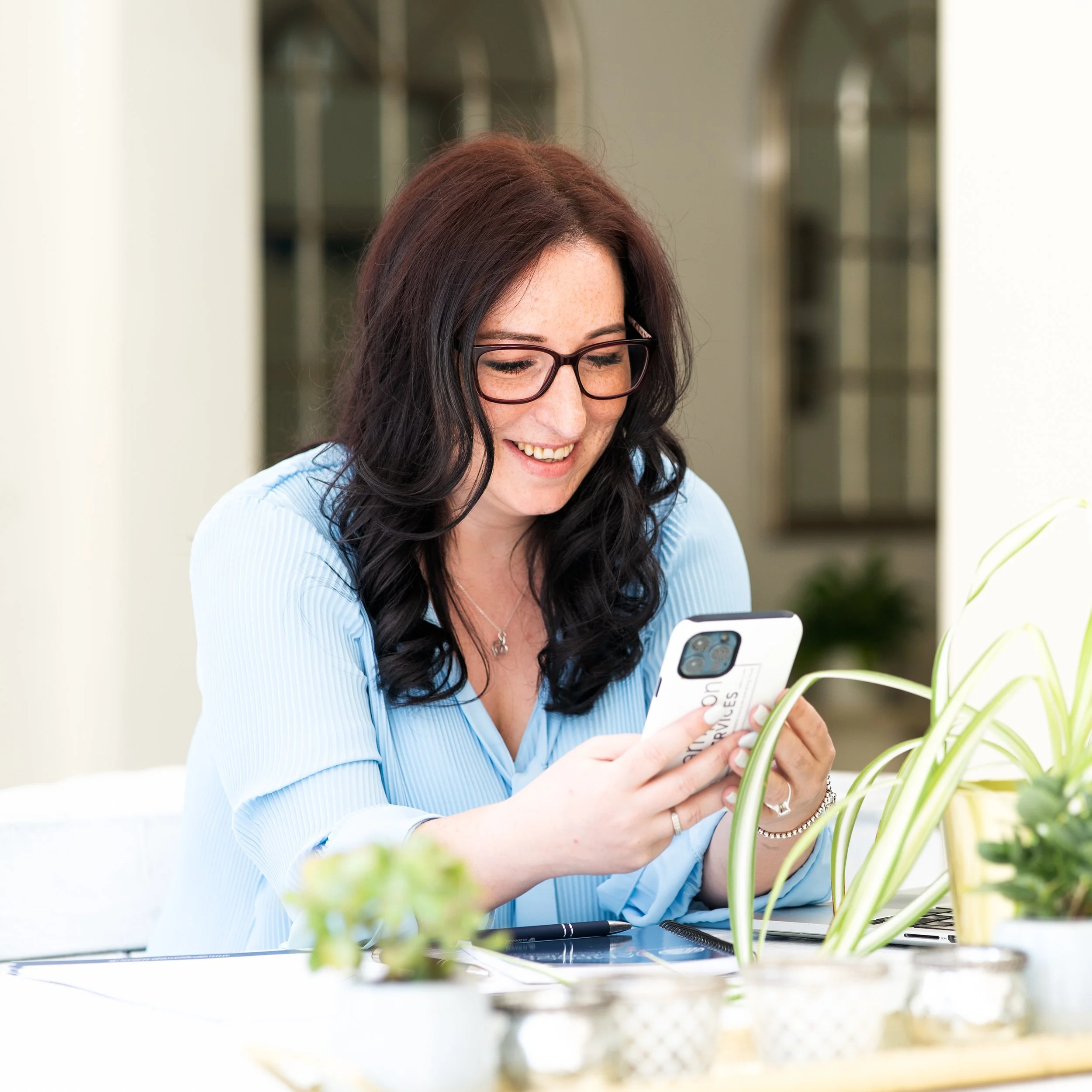 Woman in a blue blouse using her smartphone at a desk, smiling in a bright, plant-filled room.