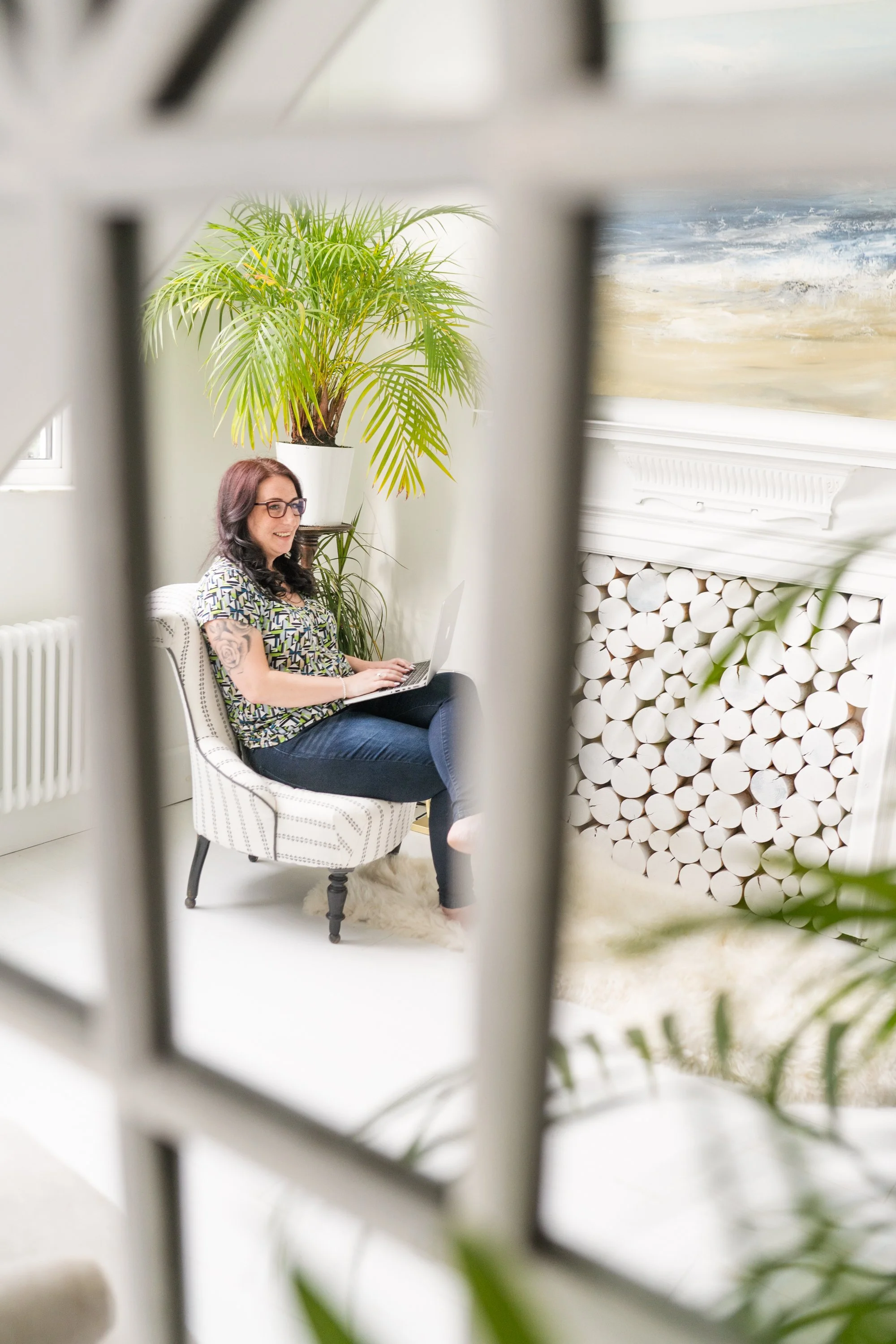 Lisa sitting in an armchair using a laptop in a bright room, viewed through a window frame, with a fireplace and large houseplant in the background.