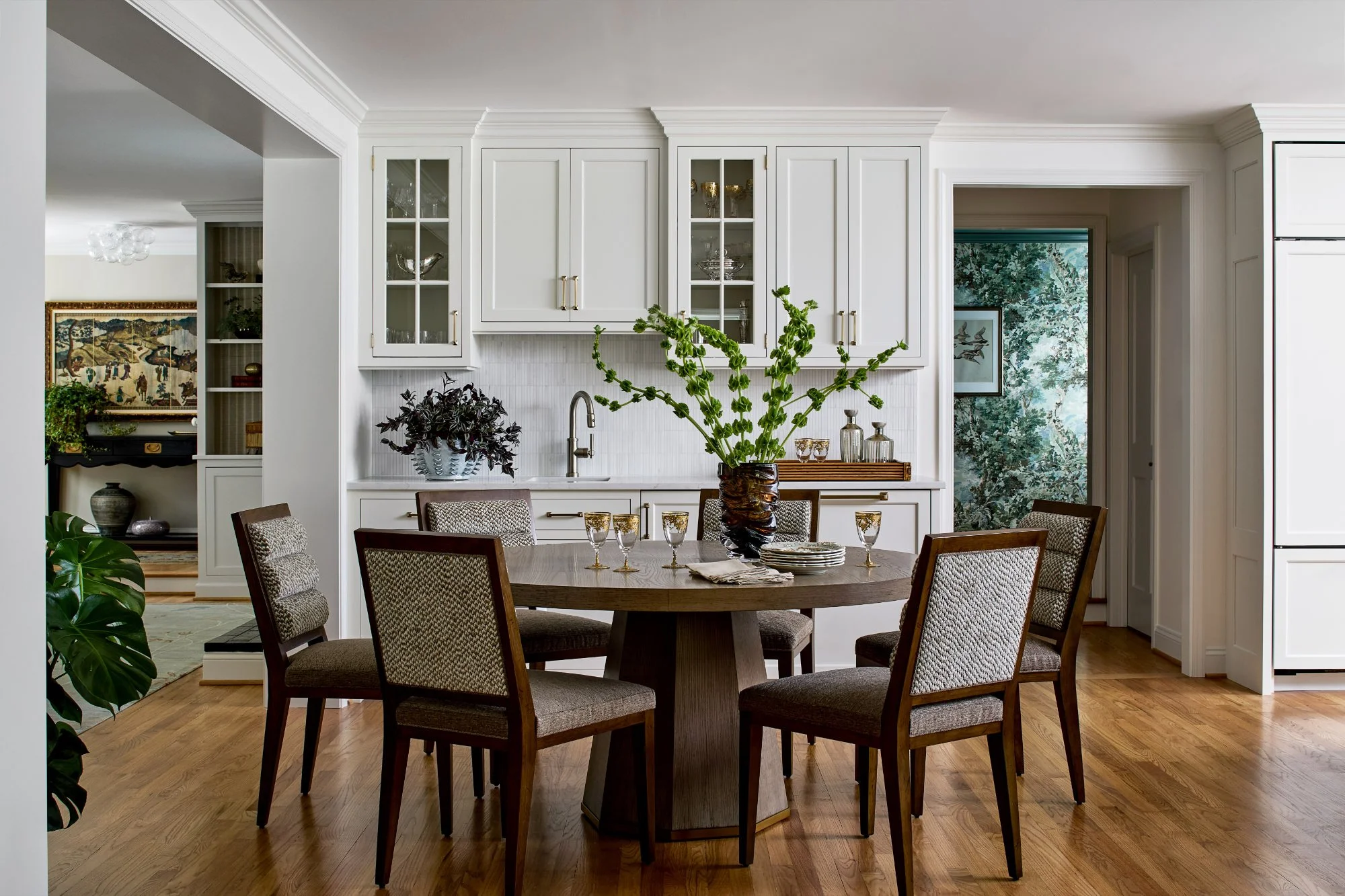 A dining area with a round wooden table, six upholstered chairs, and a large window opening into a kitchen with white cabinets and a countertop with a sink. Decorative glassware and plants are on the tabletop and shelves.