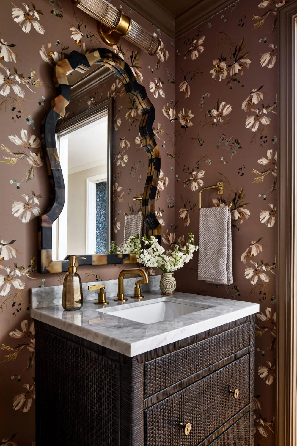 Decorative bathroom with floral wallpaper, a wavy framed mirror, marble countertop with brass fixtures, a vase of white flowers, and a towel on a brass towel holder.