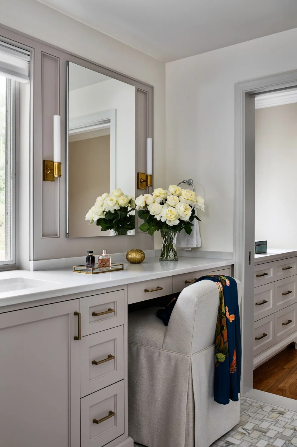Bathroom vanity with a large mirror, white flowers in a glass vase, perfume bottles, a small decorative item, a window, and a beige upholstered chair with a colorful scarf draped over the back.