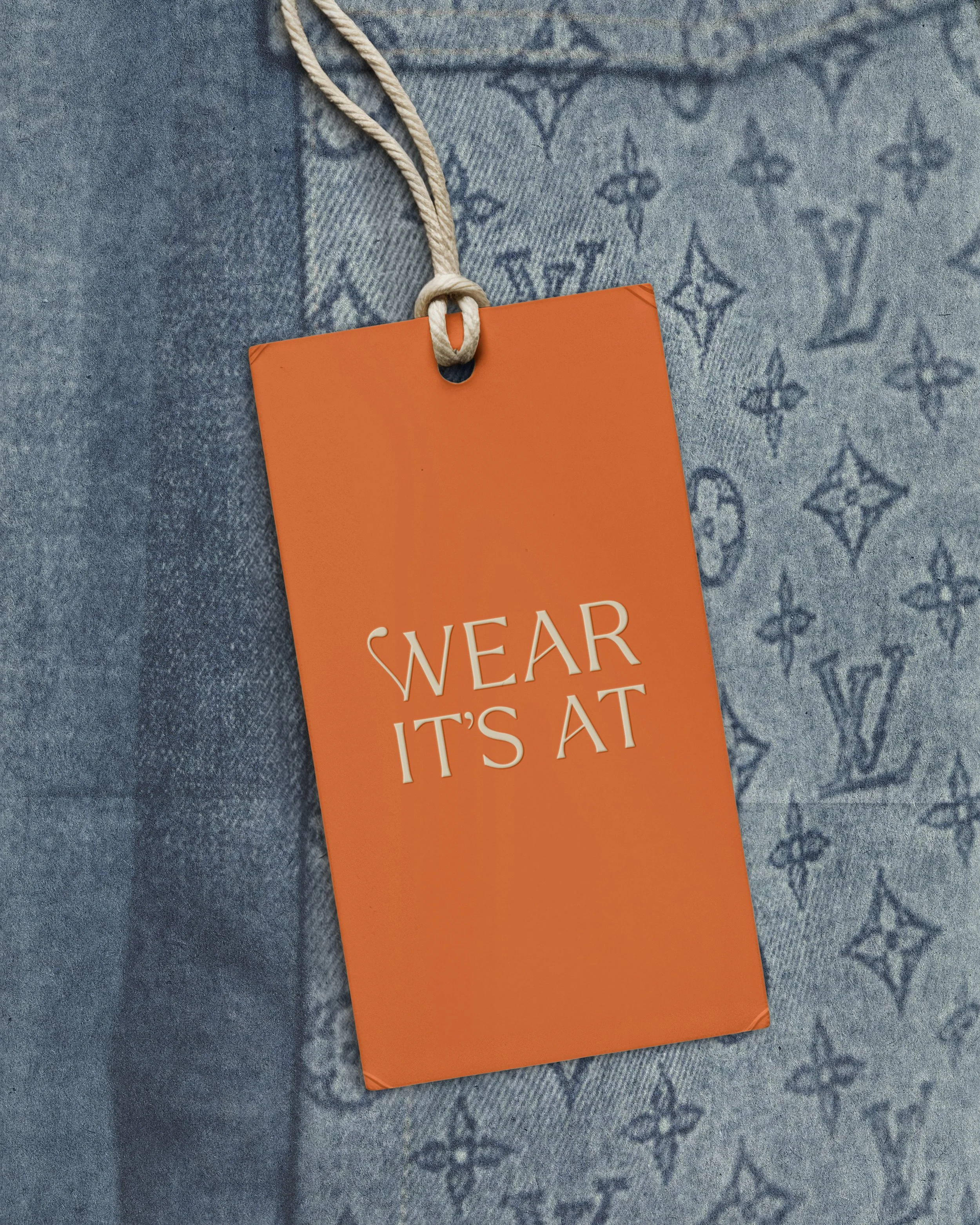 Orange clothing tag with the words 'Wear It’s At' hanging on a beige string, placed on blue fabric with a designer monogram pattern.