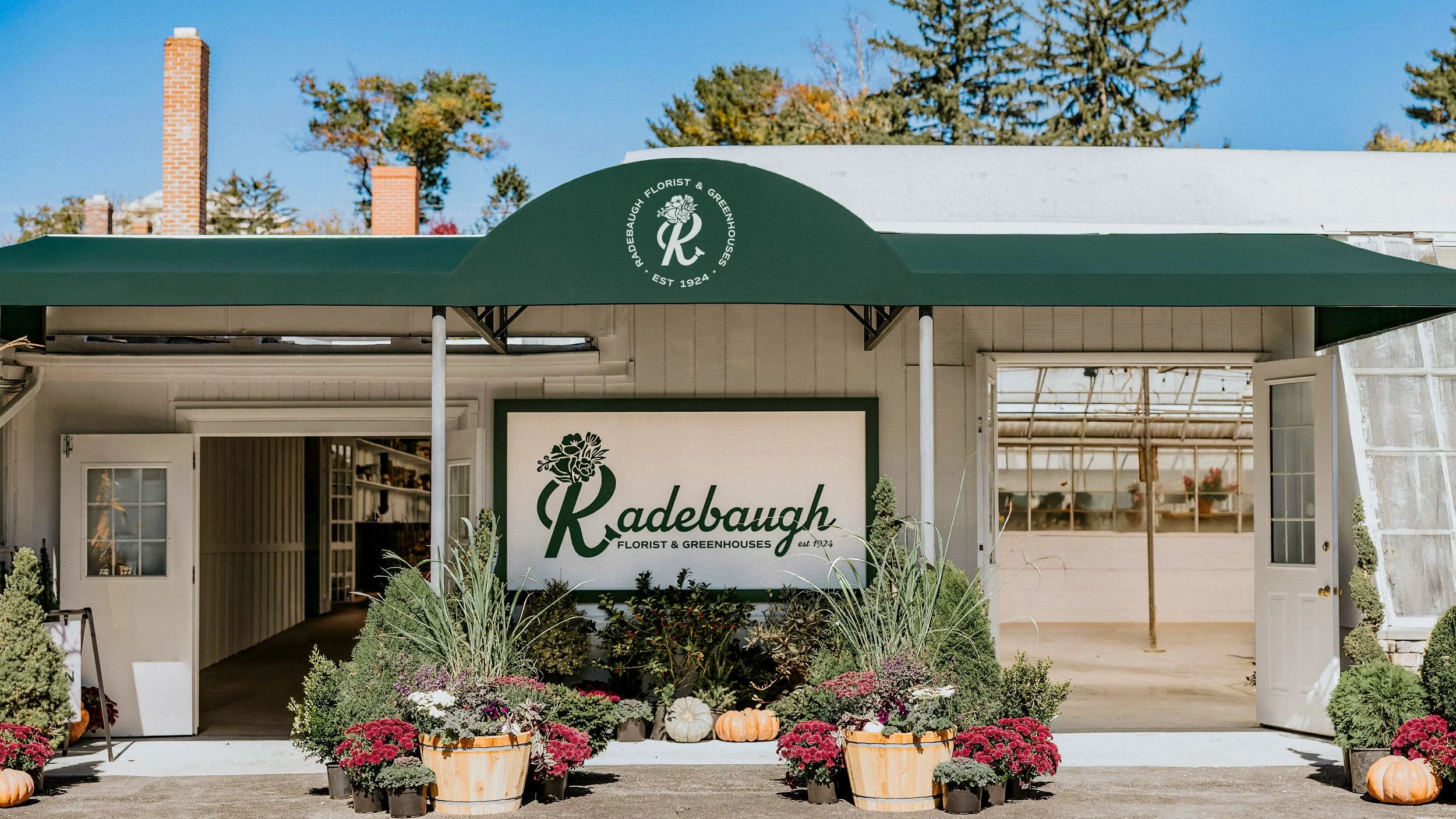 Exterior of Radebaugh Florist & Greenhouses shop with green awning and sign, surrounded by potted plants, flowers, and pumpkins during daytime.