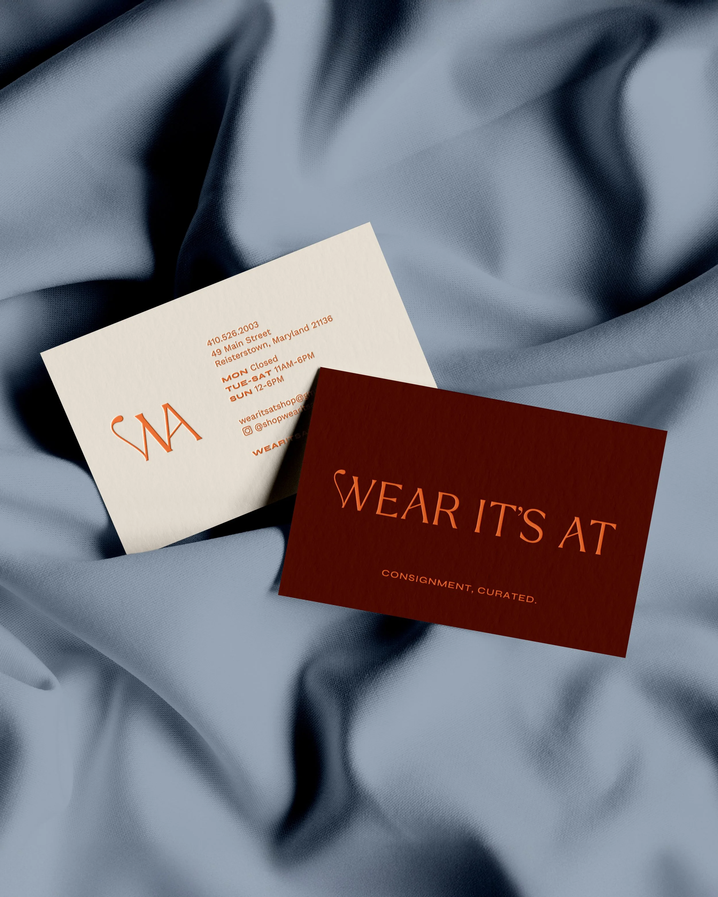 Business cards on a light blue fabric. One card is white with orange text, the other is dark red with orange text. Both promote a clothing store with the message 'Wear it’s at'.
